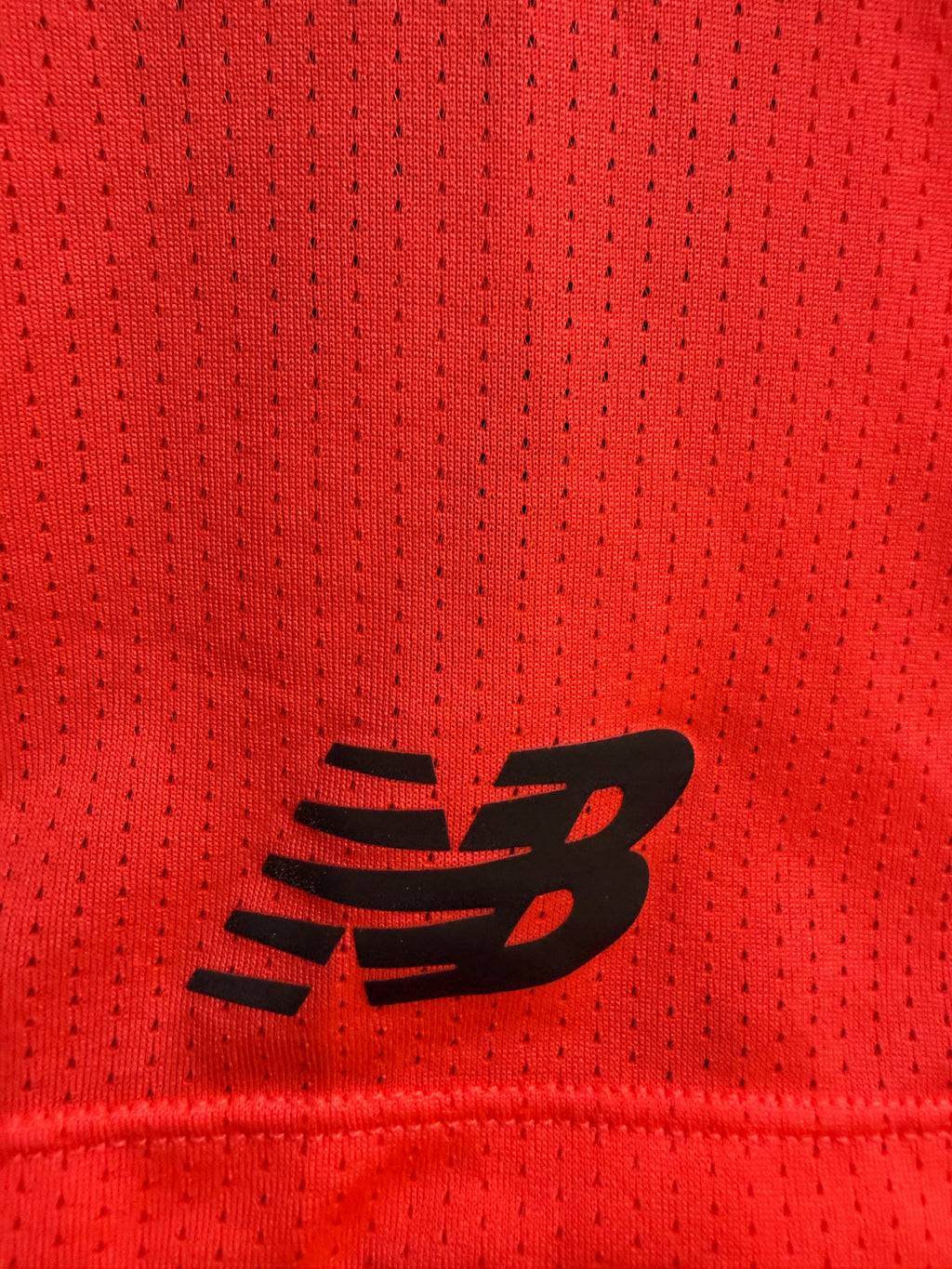 Close-up of red sports T-shirt showing black New Balance logo on breathable mesh fabric