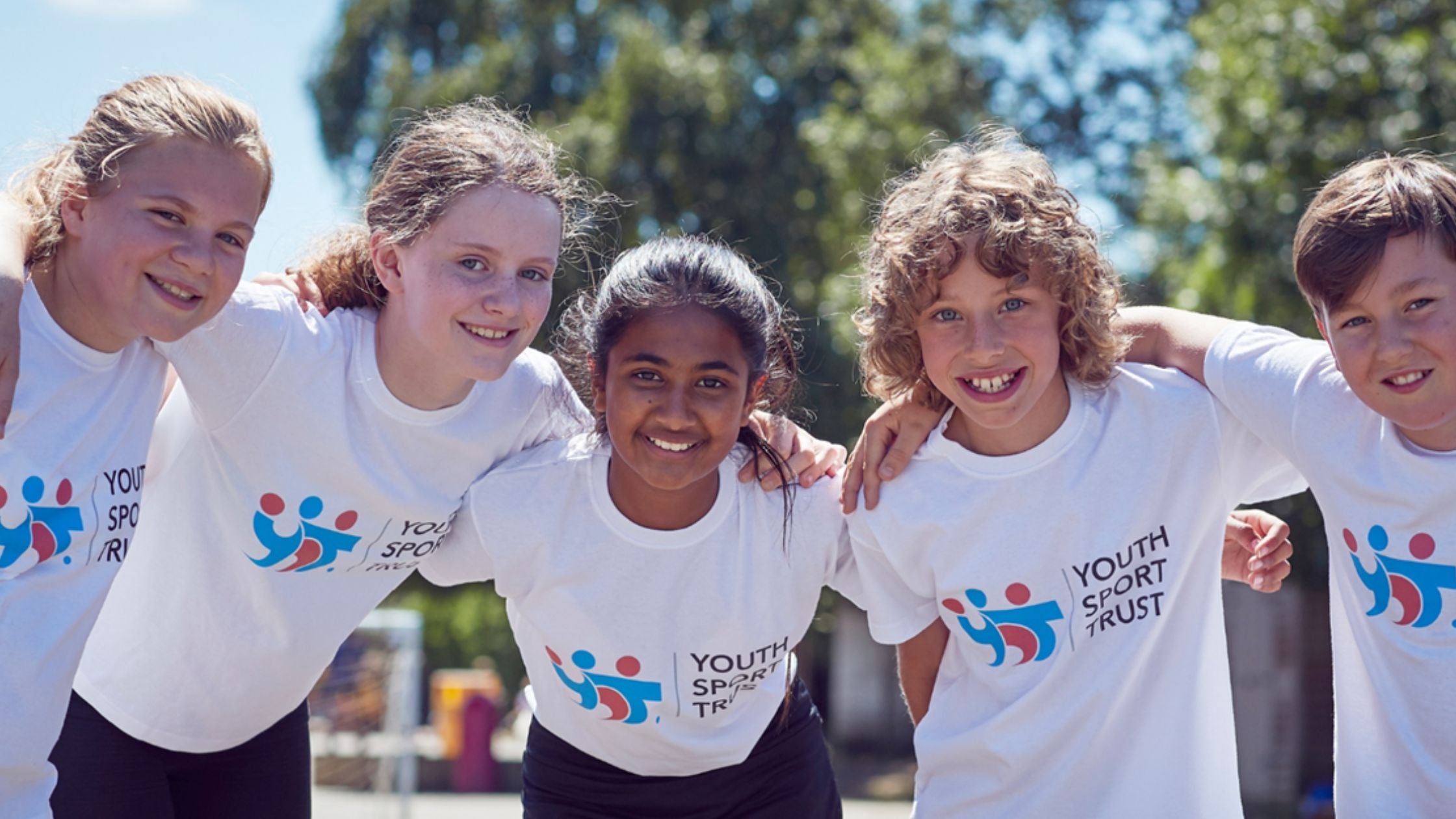 Five children wearing 'Youth Sport Trust' t-shirts, smiling and posing together outdoors.