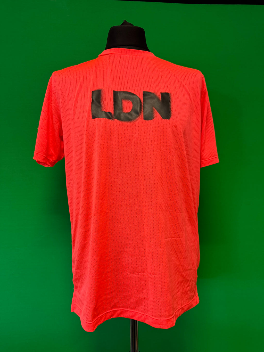 Red sports T-shirt with black LDN lettering on the back displayed on a mannequin against green background