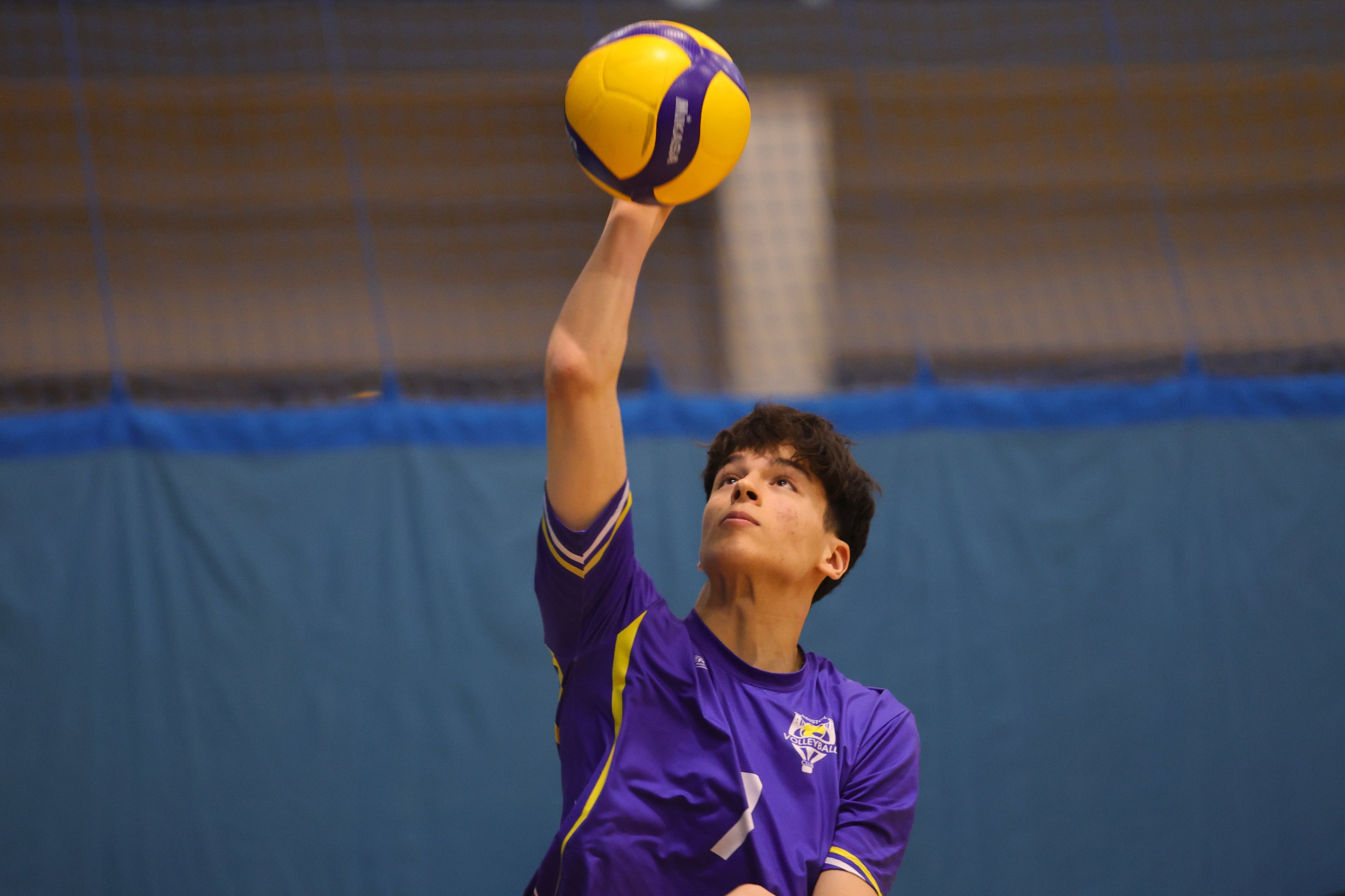Person in purple volleyball uniform preparing to serve a yellow and purple volleyball indoors.