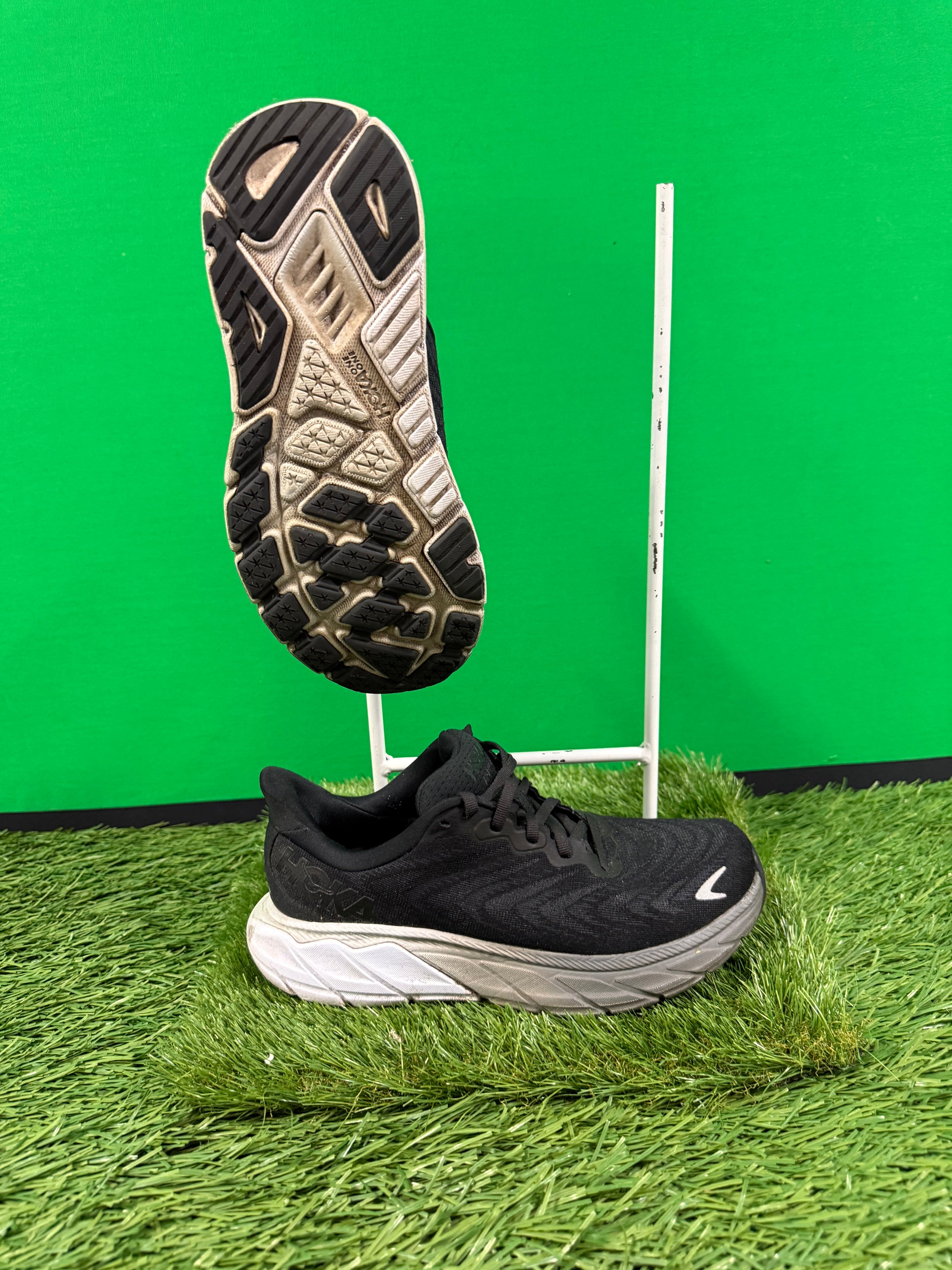 Side and sole view of black Hoka running trainers displayed against green background on artificial grass