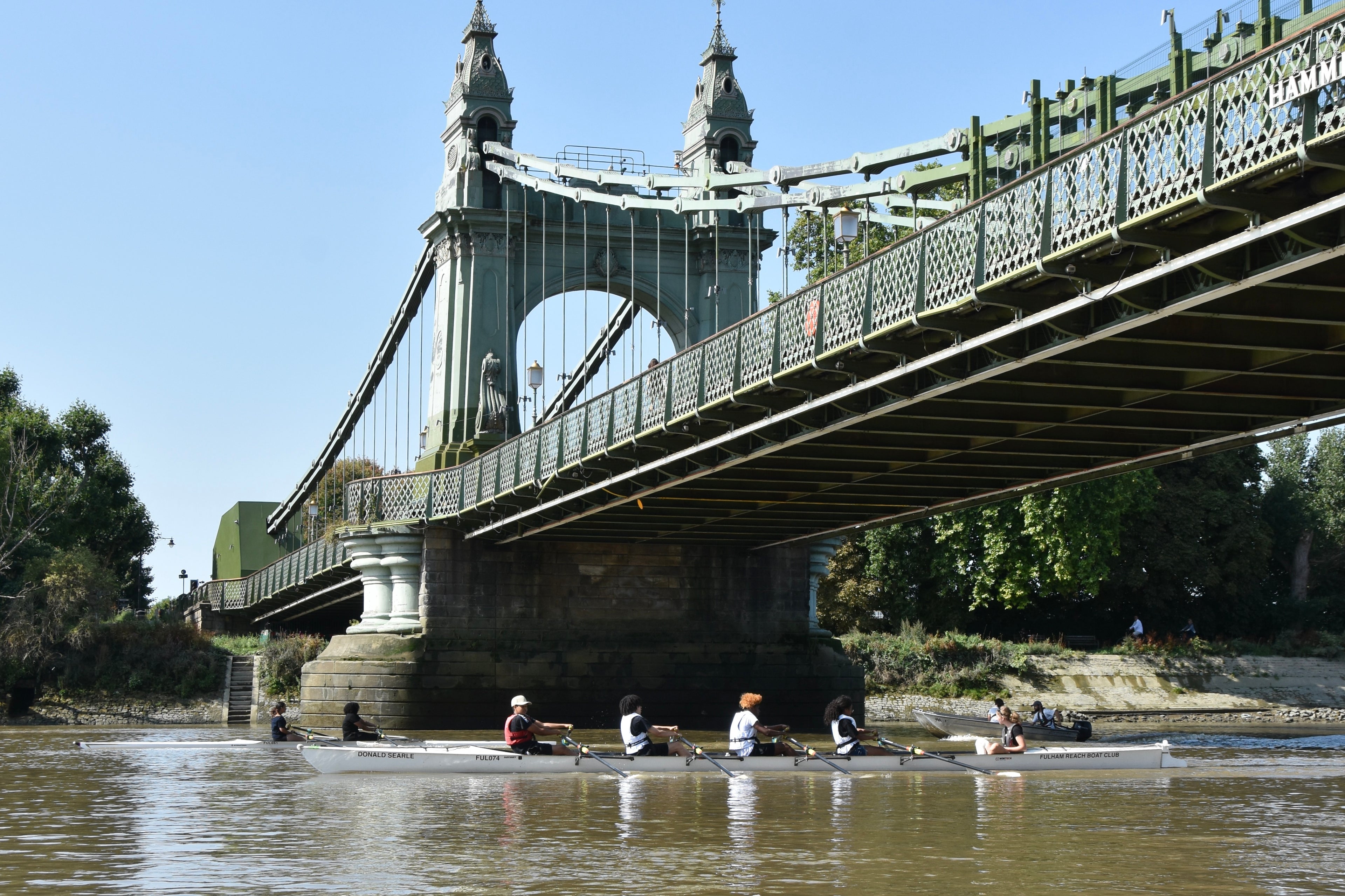 Rowers on a boat under a large green bridge with a clear blue sky.