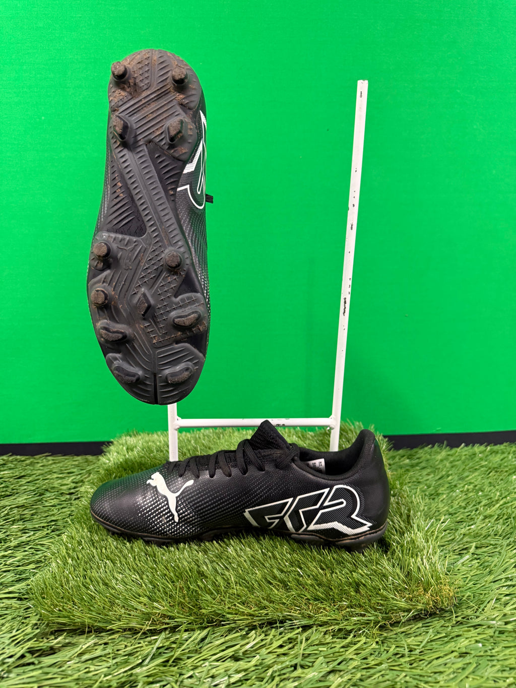 Side view of black Puma football boots on artificial grass with studded sole visible on suspended boot against green background