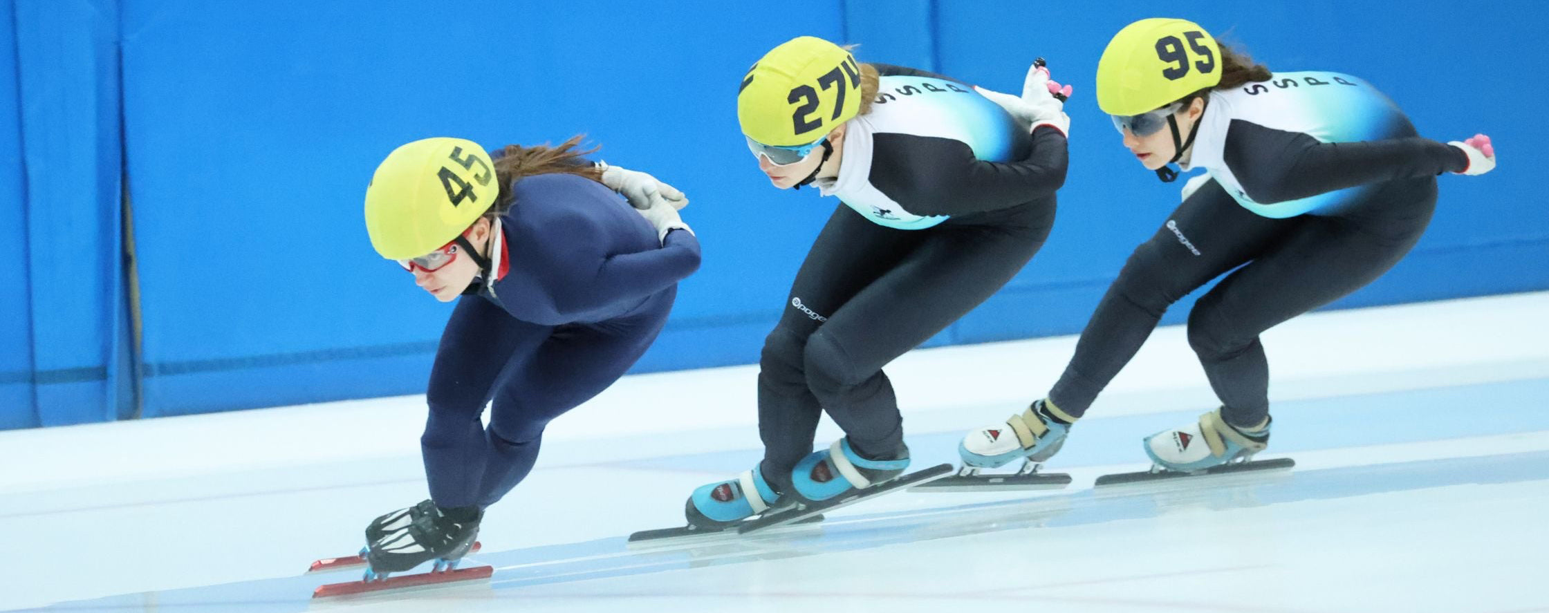 Three speed skaters in a race on an ice track with blue walls in the background.