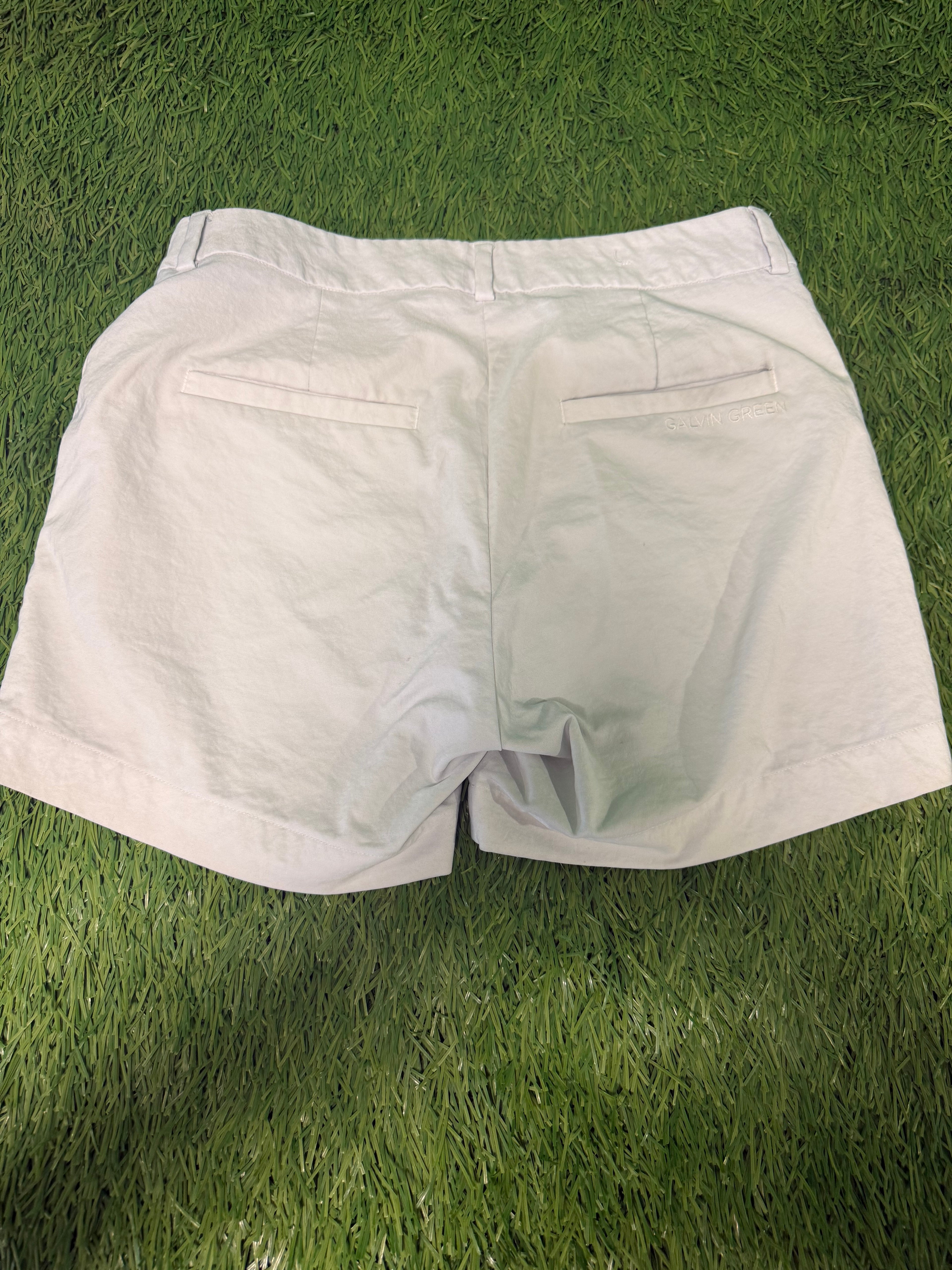 Back view of white women's golf shorts laid flat on grass, showing double welt pockets and brand embroidery