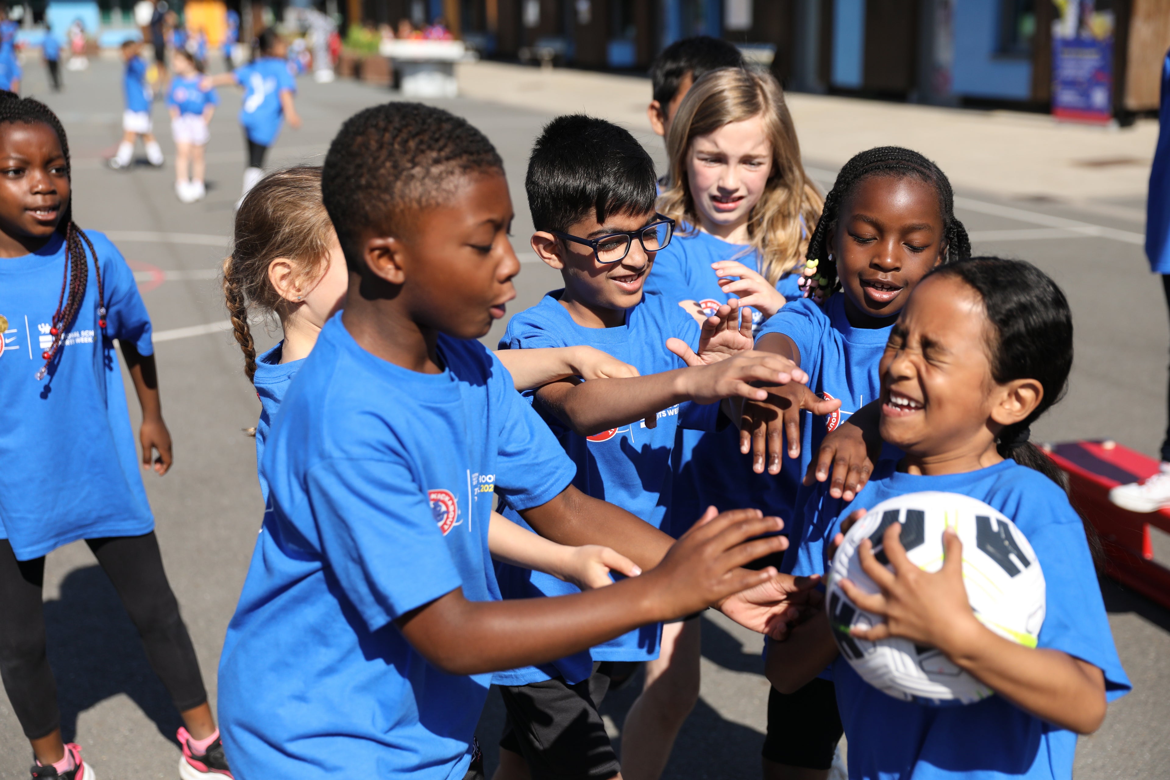 Children in blue shirts playing with a soccer ball on a street.