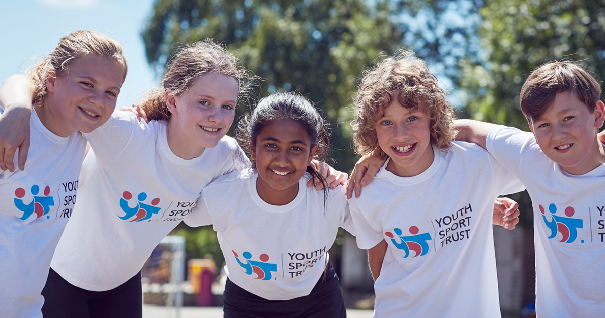 Five children wearing white shirts with a logo, standing outdoors on a sunny day.
