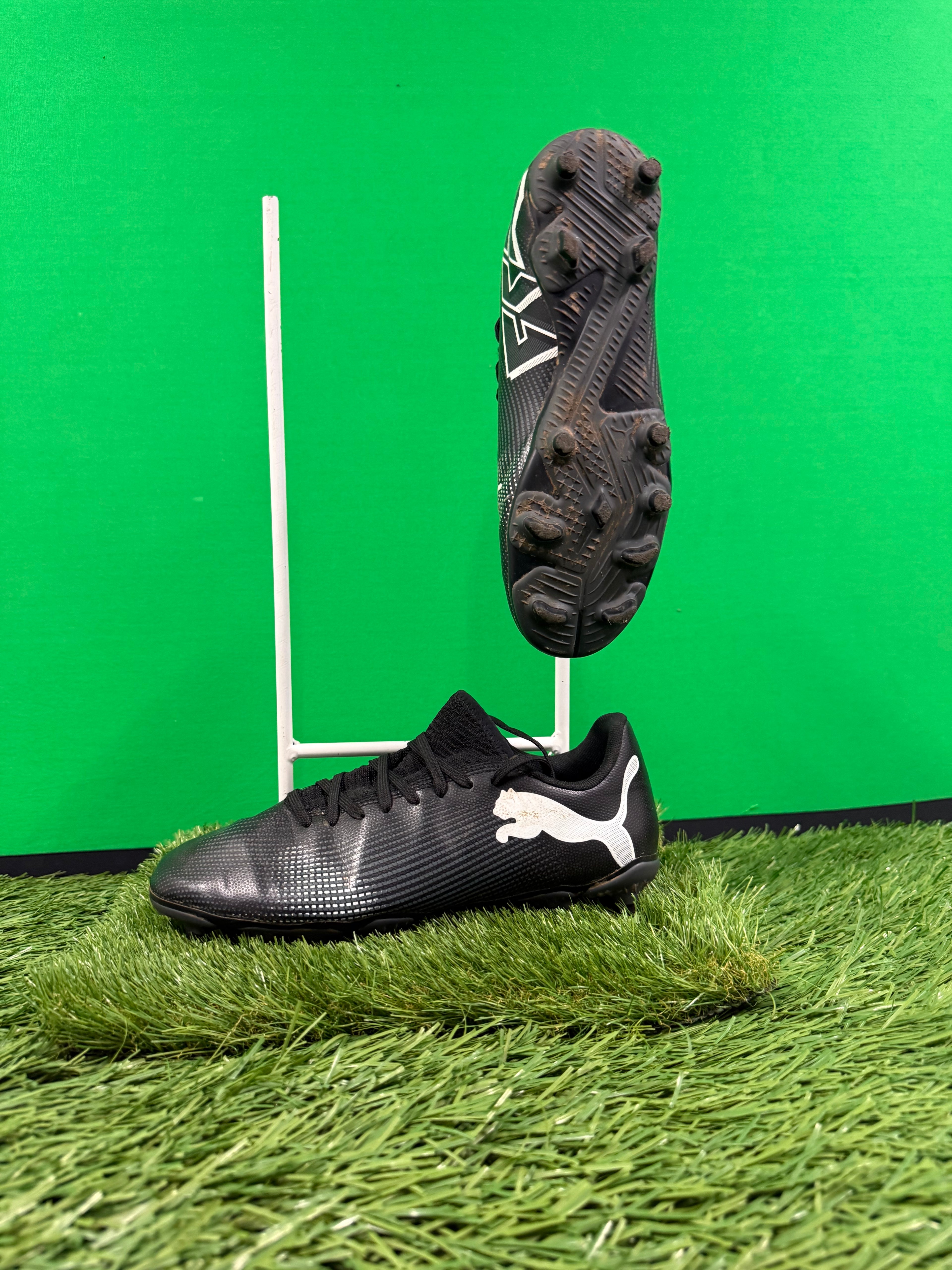 Pair of black Puma football boots on artificial grass with one boot resting on ground and the other suspended showing studded sole