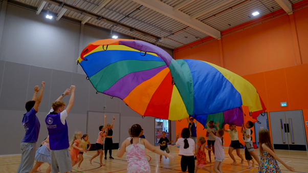 Children playing with a large colorful parachute in an indoor gymnasium.