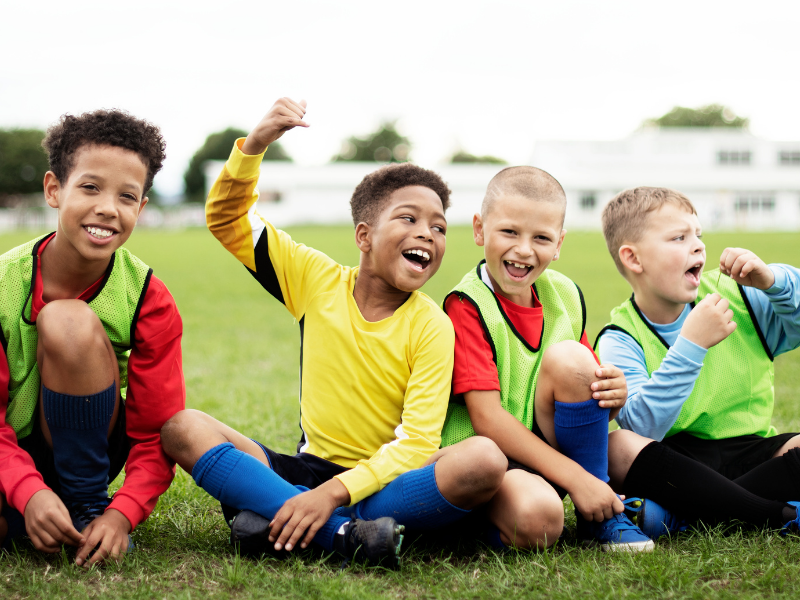 Four children in sports attire sitting on a grassy field.