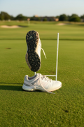 Pair of white Adidas golf shoes on artificial grass, one upright and one showing textured sole with grip pattern