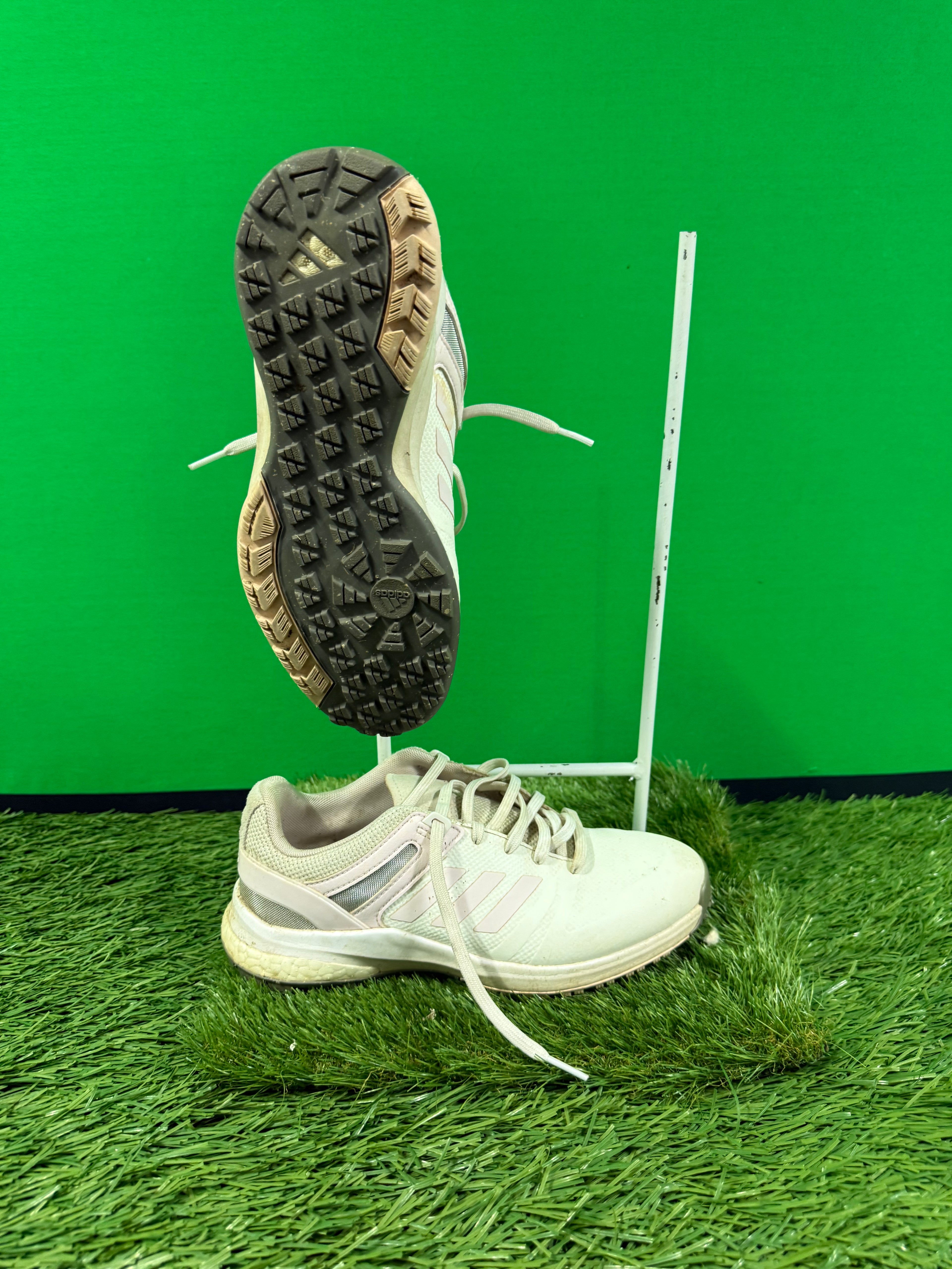 Pair of white Adidas golf shoes on artificial grass, one upright and one showing textured sole with grip pattern