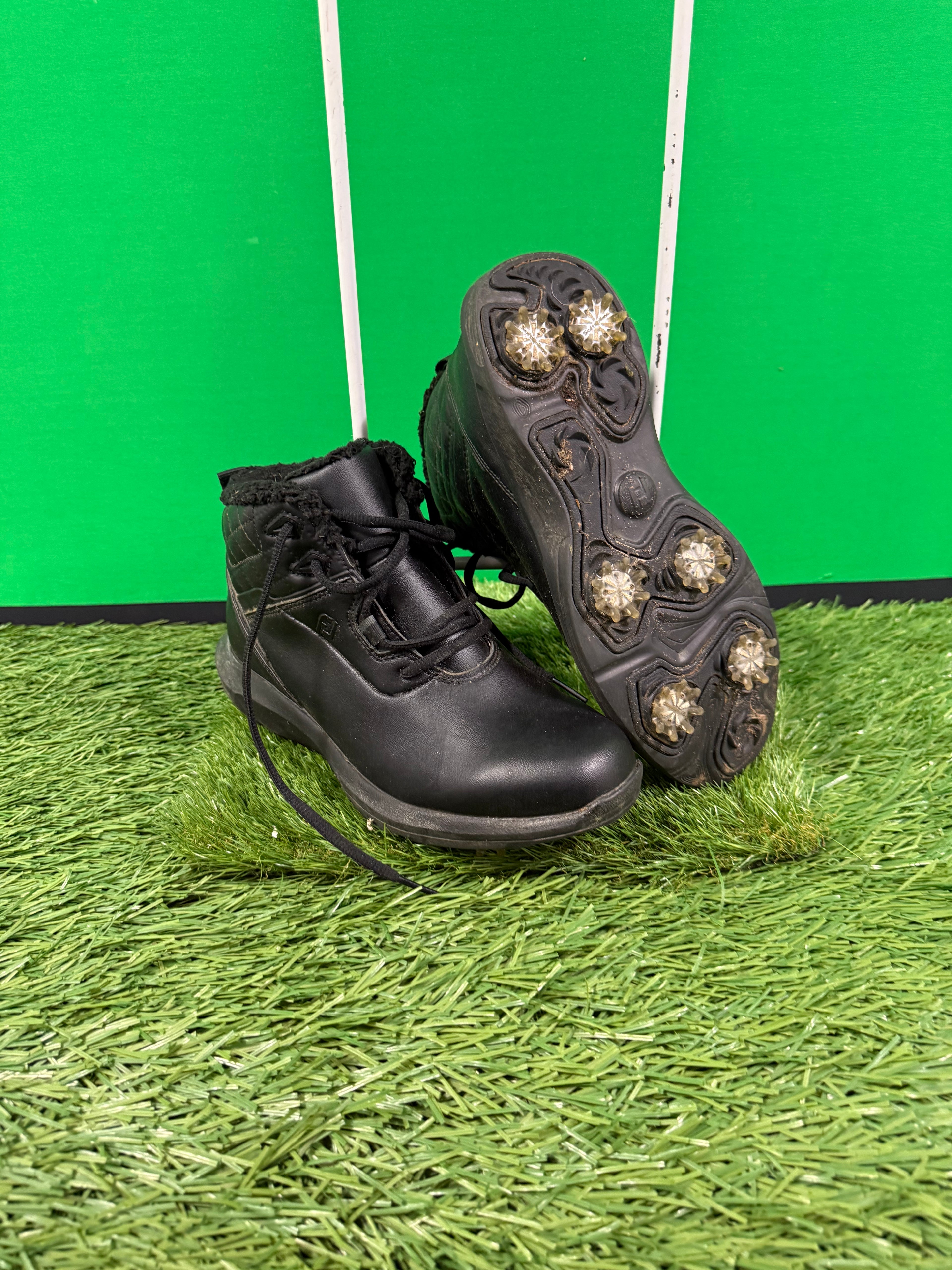 Pair of black Footjoy golf boots on artificial green grass with visible metal spikes on the sole