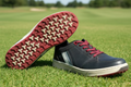black casual outdoor shoe with maroon laces and patterned rubber sole against green background