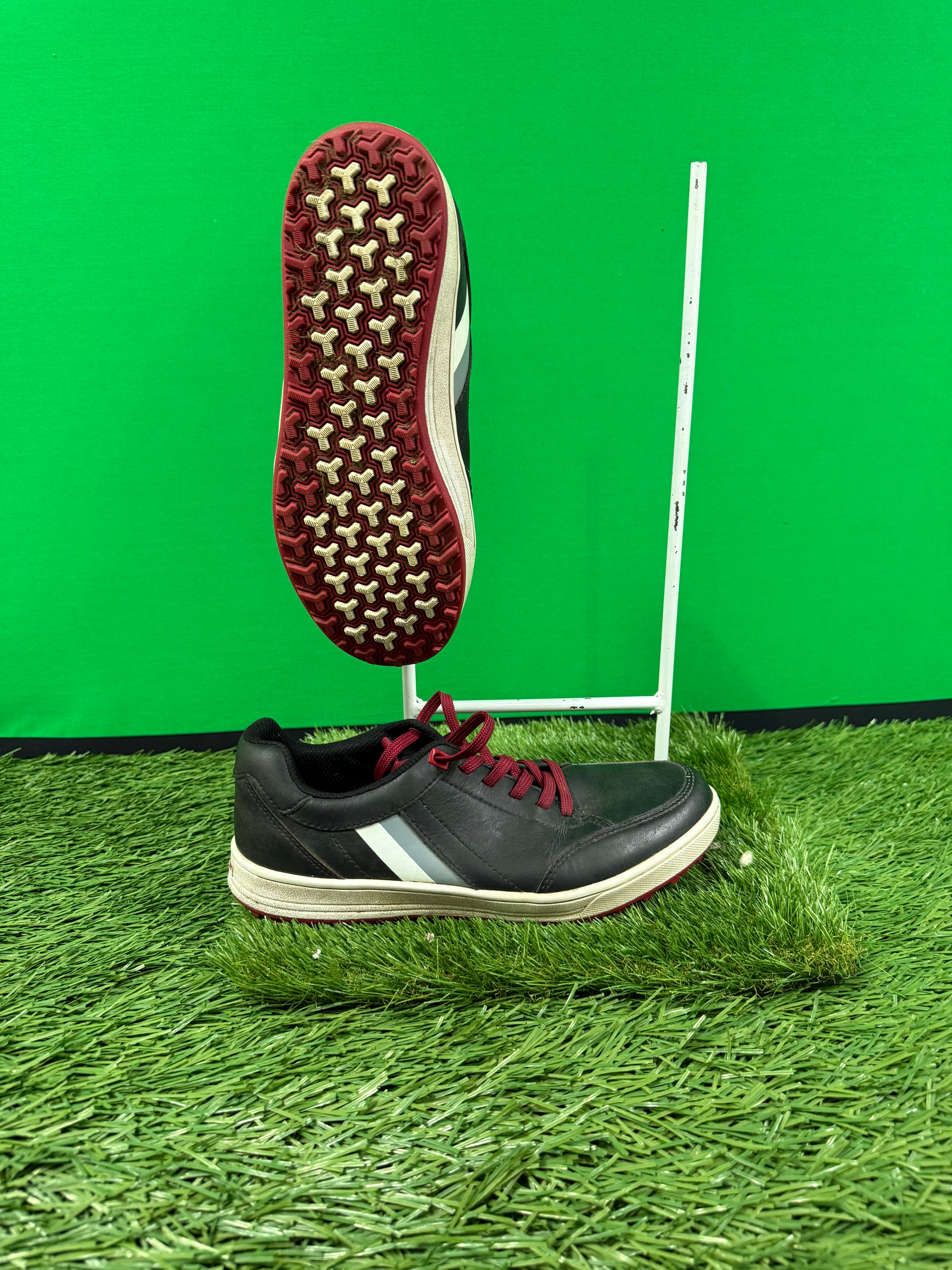 black casual outdoor shoe with maroon laces and patterned rubber sole against green background