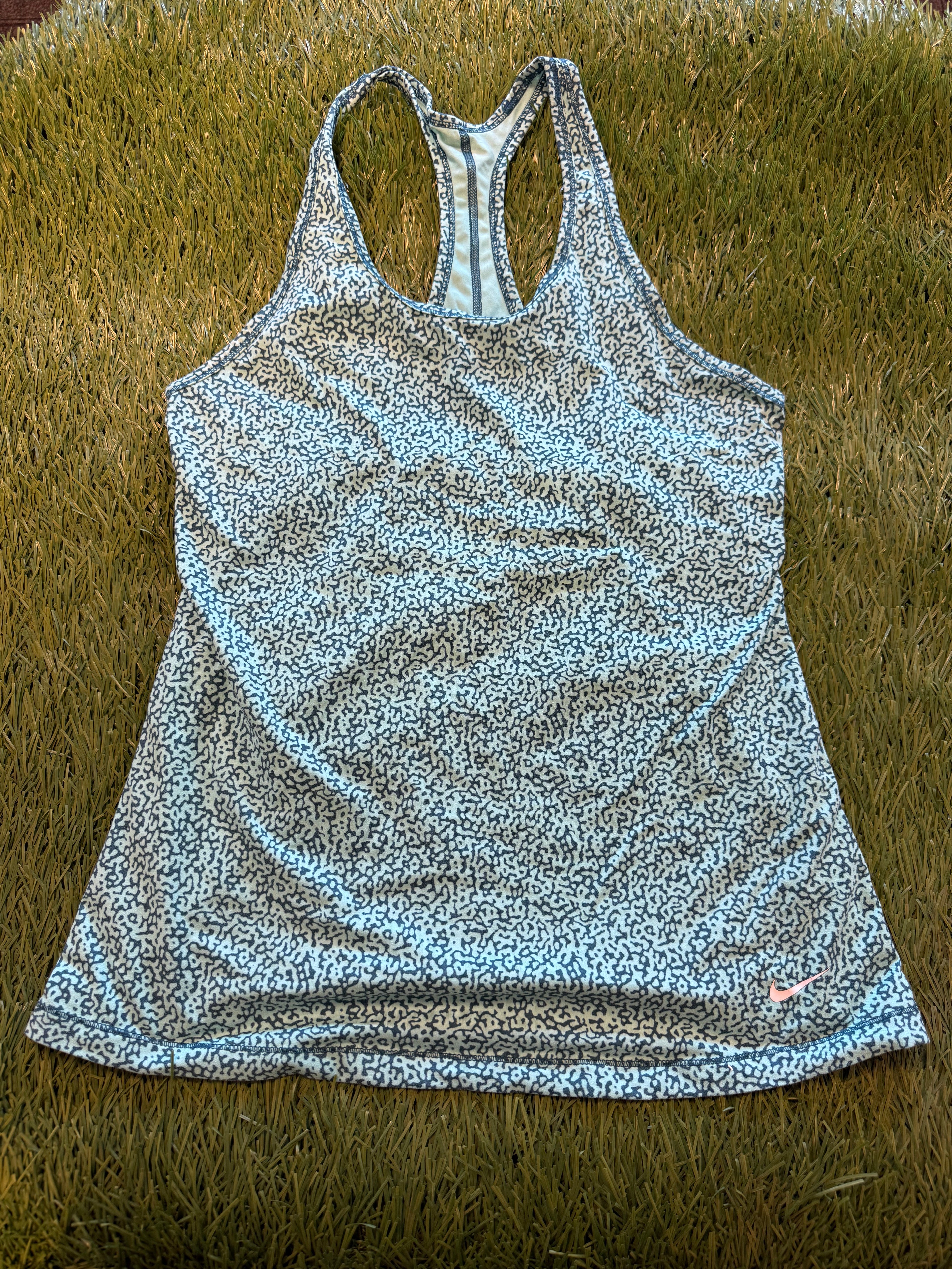 Front view of Nike women's racerback tank top with blue and white abstract pattern, laid flat on grass background, featuring pink Nike swoosh on hem.