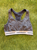 Grey New Balance sports bra laid flat on grass showing front view with branded elastic band