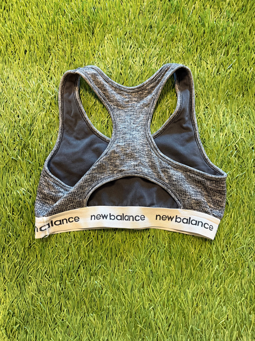 Grey New Balance sports bra laid flat on grass showing back view with racerback design and branded elastic band