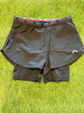 Black RUN women's running shorts with dual-layer design, elastic waistband with pink and black logo, on green grass background