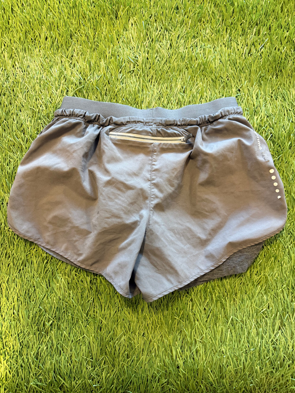Back view of grey Karrimor running shorts showing zip pocket and reflective logo, laid flat on grass