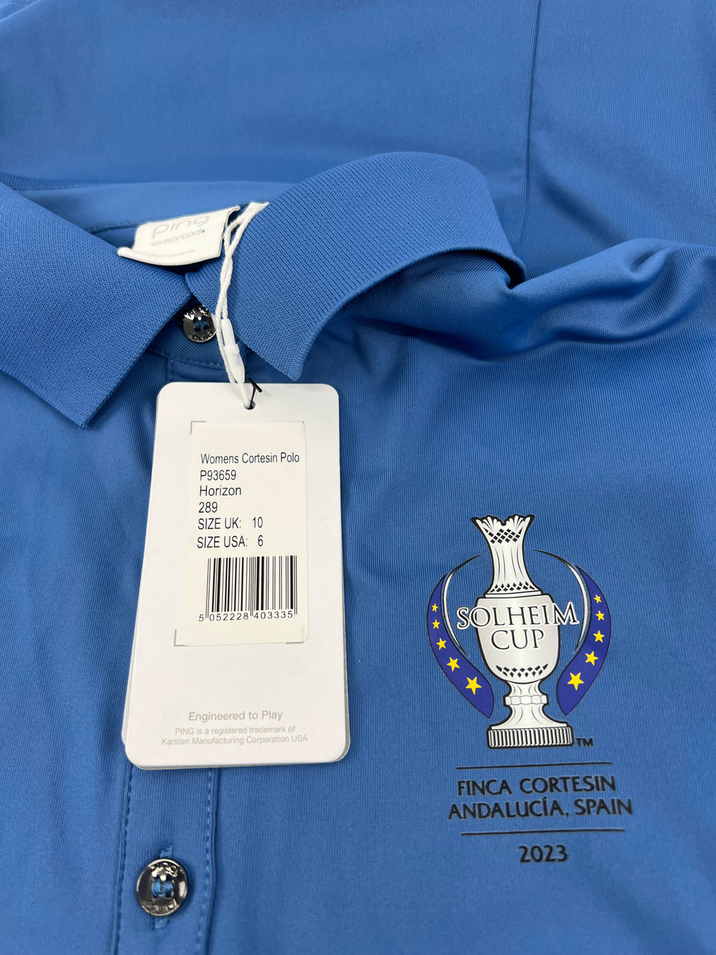 Close-up of Ping Solheim Cup 2023 embroidery and product tag detailing size and event information on horizon blue polo shirt