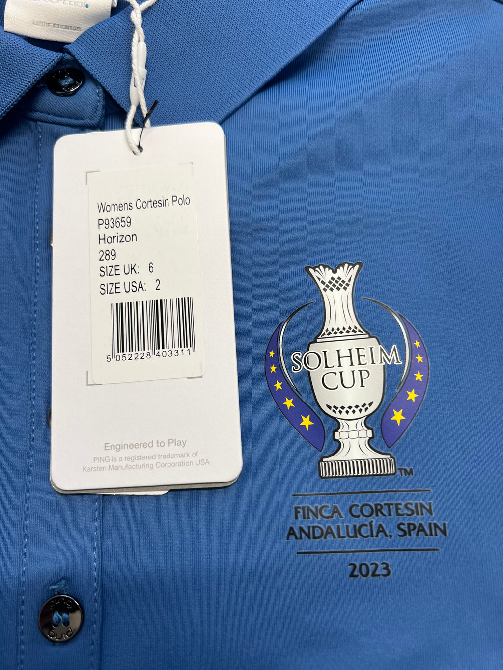 Close-up of Solheim Cup logo and product tag on blue Women's Ping polo shirt