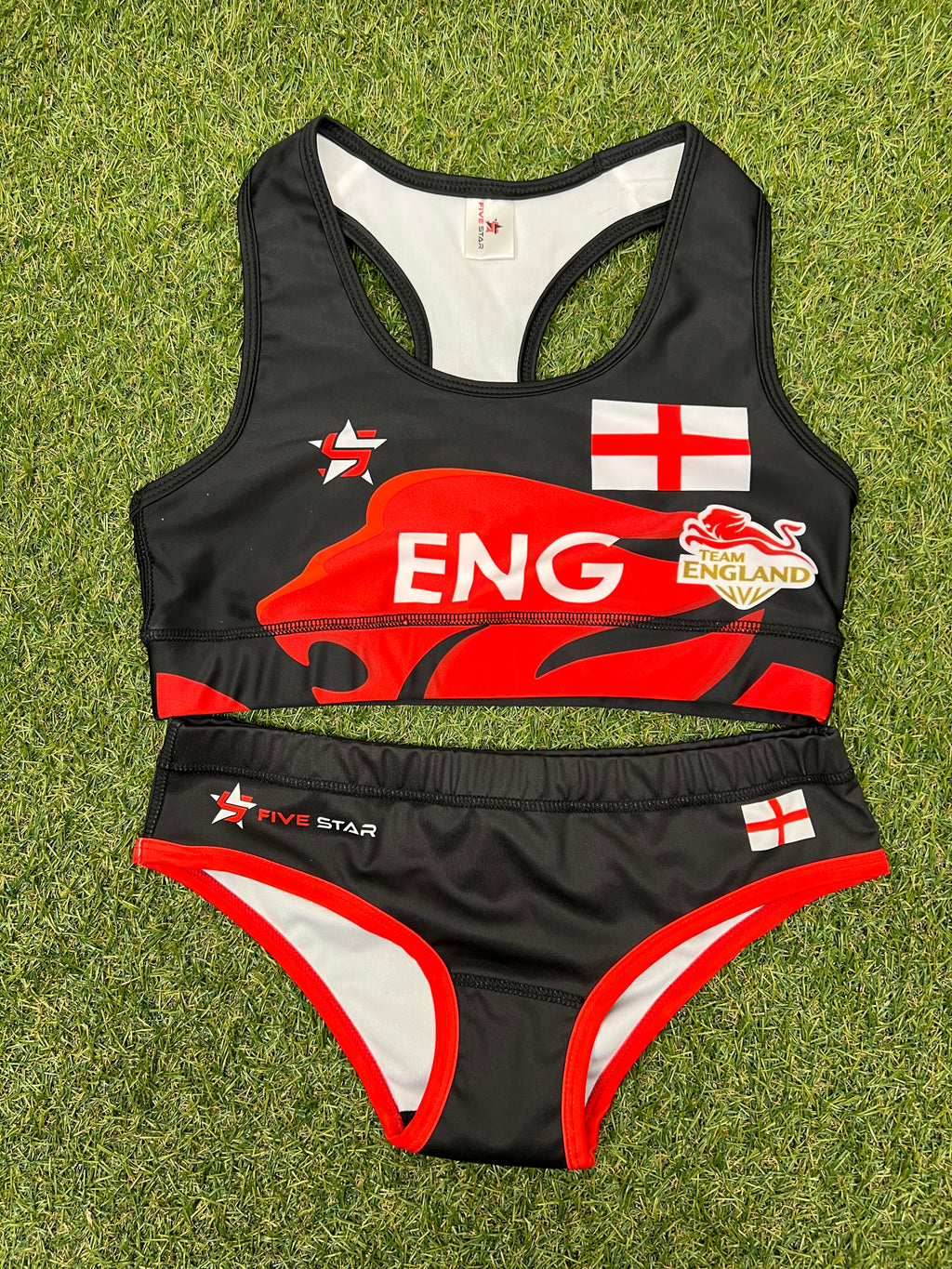 Front view of women’s Team England black and red beach volleyball bikini laid out on grass