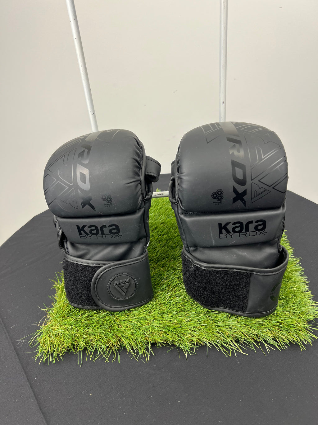 Front view of Kara by RDX 7oz black grappling gloves with velvet wrist straps on artificial grass