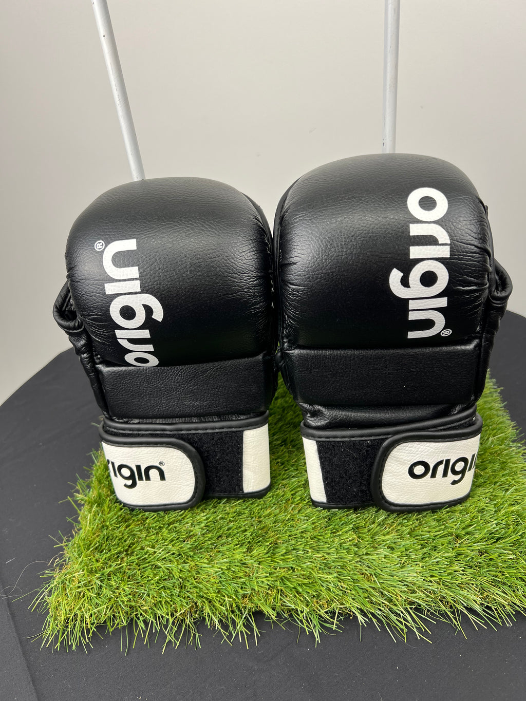 Front view of black Origin grappling gloves with white wrist straps placed on artificial grass