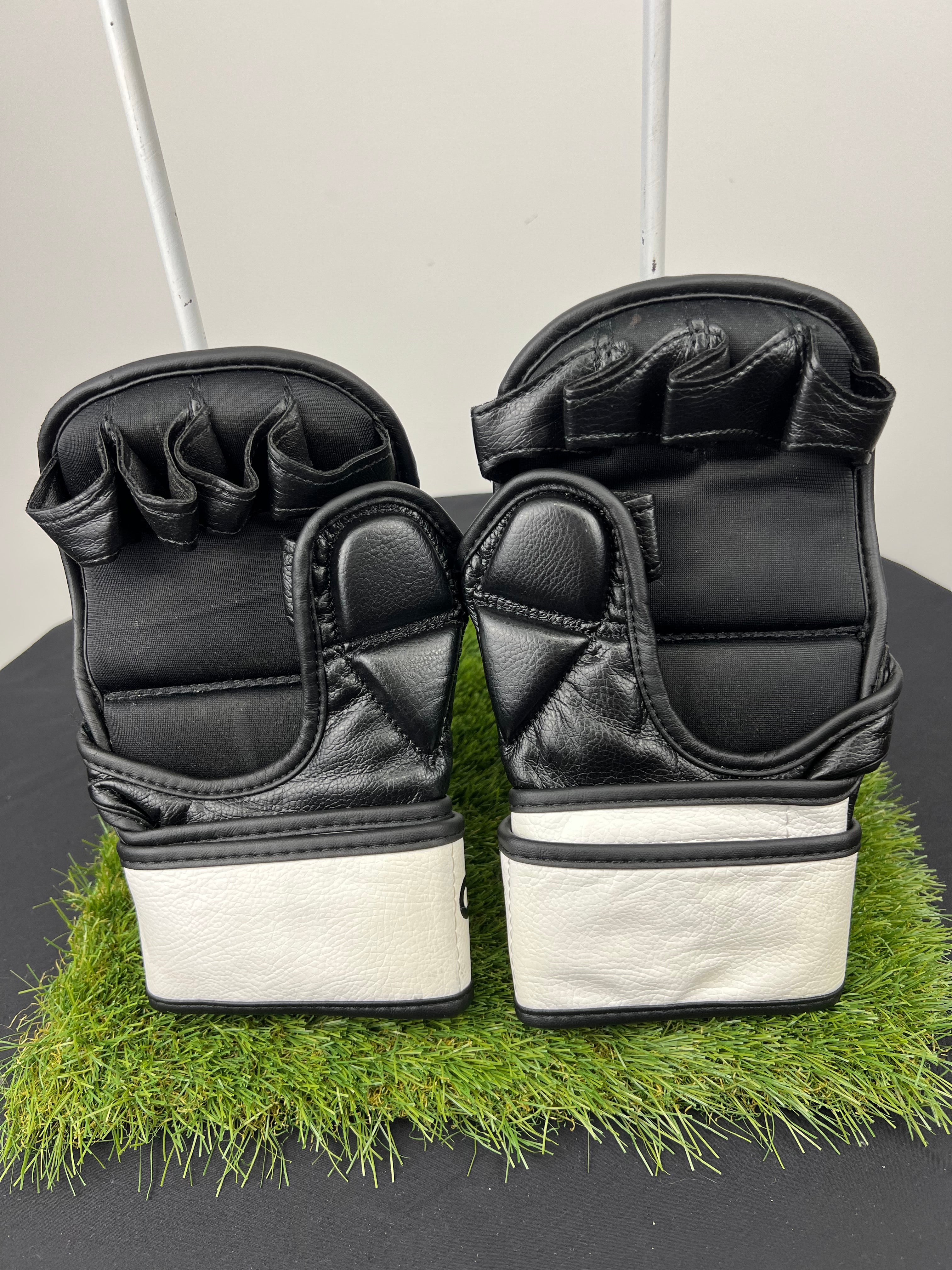 Back view of black Origin grappling gloves showing finger slots and white adjustable wrist straps on artificial grass