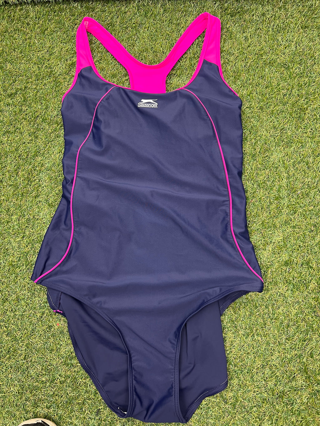 Front view of women's Slazenger racer back swimsuit in purple with pink straps and piping, laid flat on grass