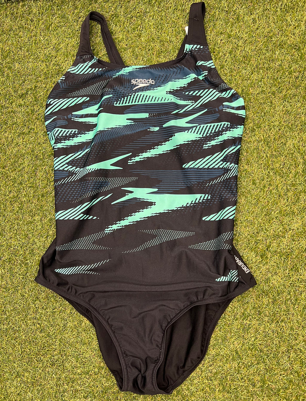 Front view of women’s Speedo swimsuit in black with mint and navy abstract pattern, laid flat on grass