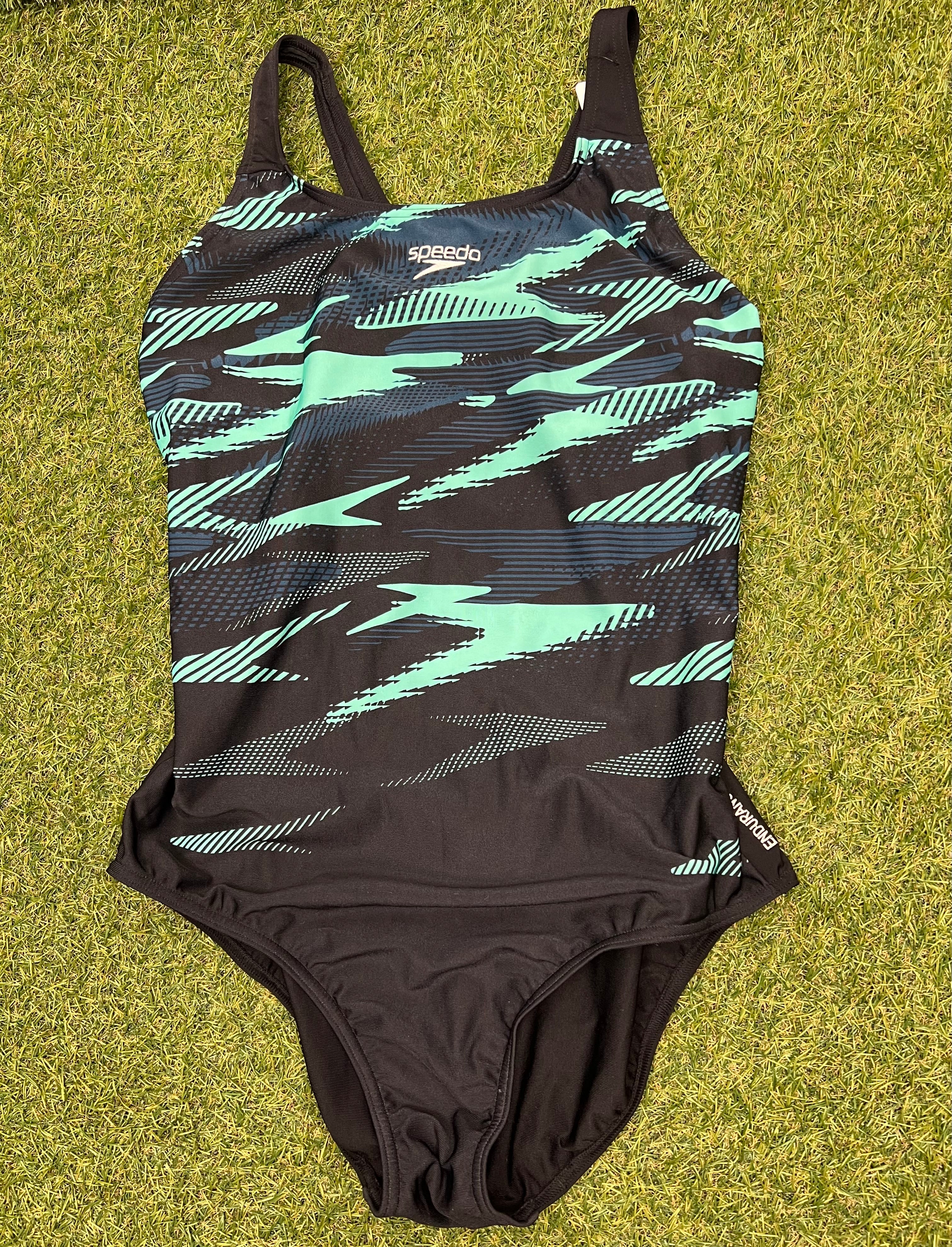 Front view of women’s Speedo swimsuit in black with mint and navy abstract pattern, laid flat on grass