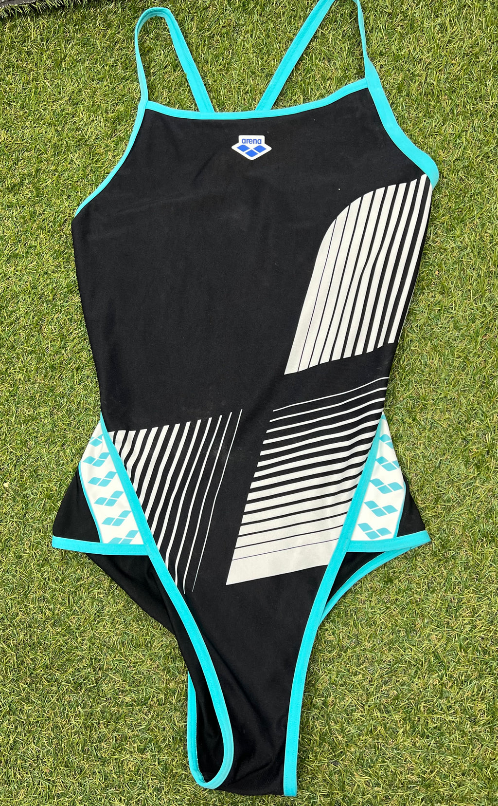 Front view of women’s black arena swimsuit with white geometric stripes and turquoise trim laid on grass