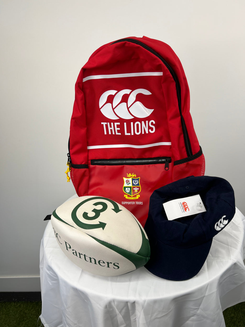 Red rugby supporters backpack with The Lions logo, crest, and zipper pocket with a white and green rugby ball and navy Canterbury cap on white table