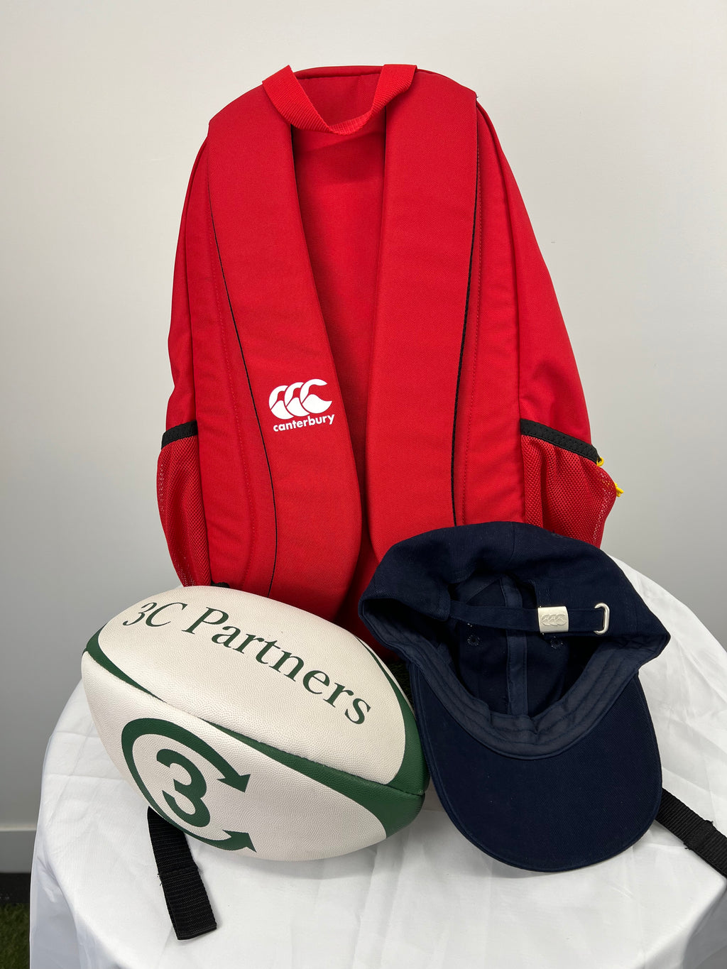 Back view of red rugby supporters backpack showing padded straps and black mesh side pockets alongside white and green rugby ball and navy Canterbury cap