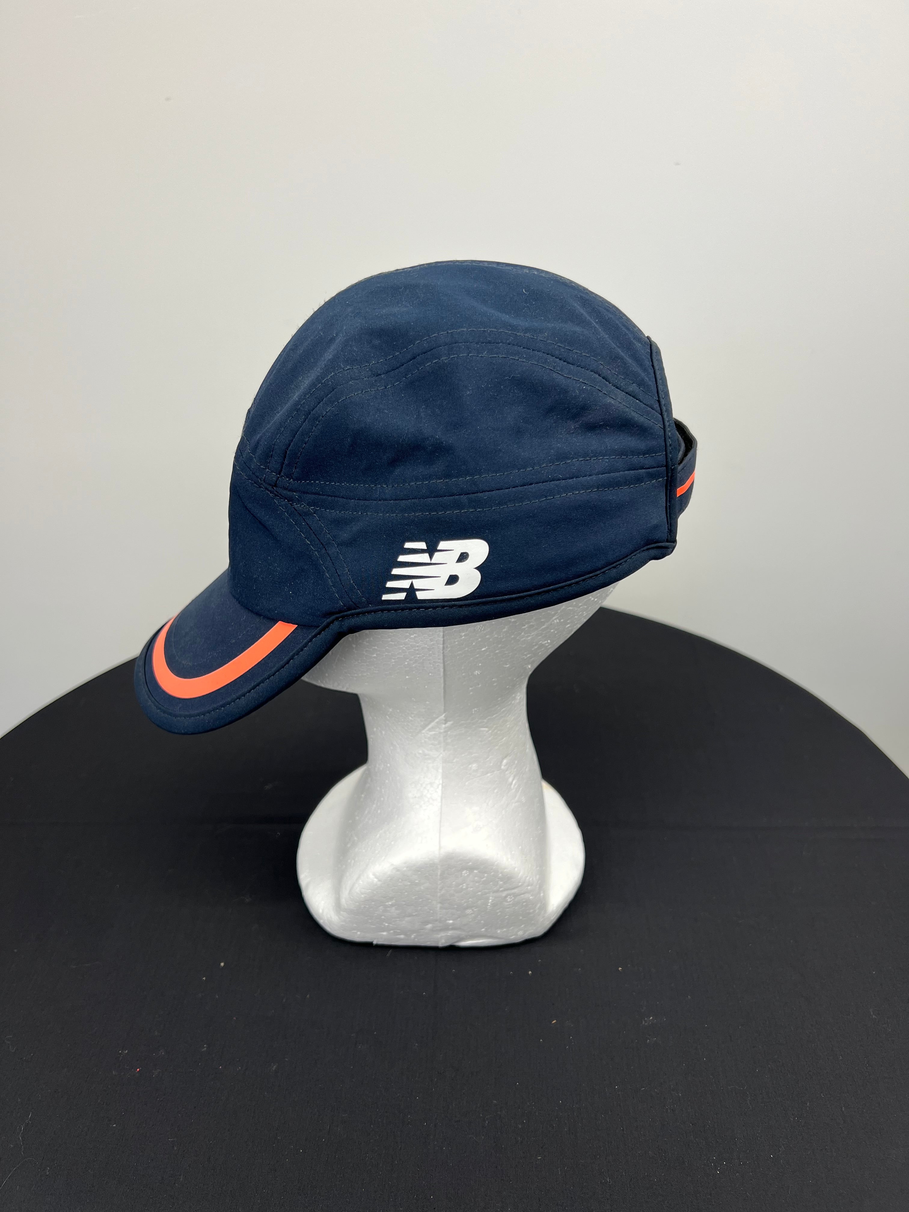 Side view showing New Balance NB logo on navy cricket baseball cap with orange-trimmed visor