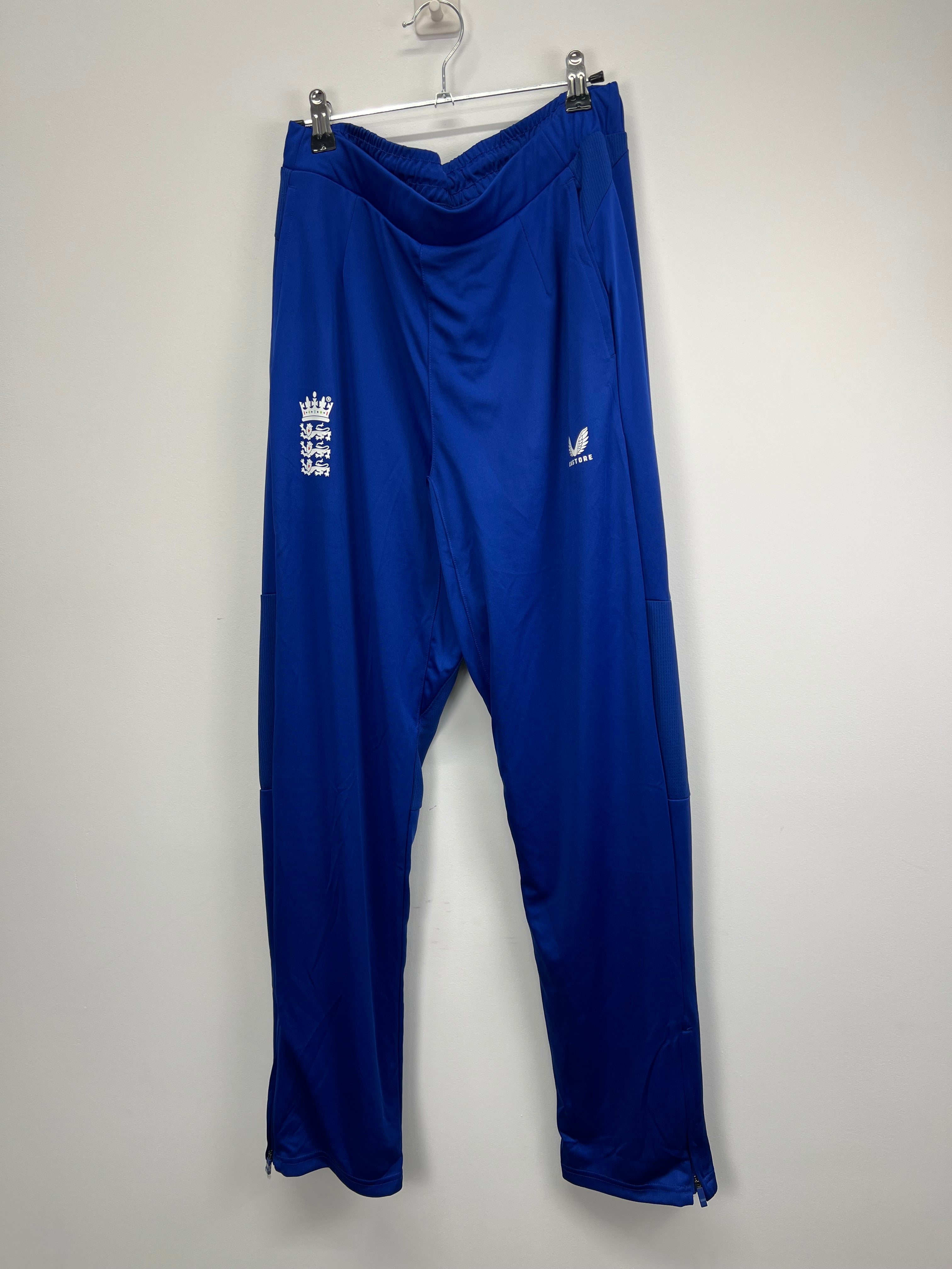 Men’s Castore England cricket tracksuit bottoms in royal blue hanging on a hanger, front view showing logos