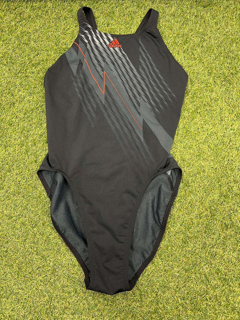 Front view of Adidas Infinitex black swimsuit with grey and orange graphic design on green grass
