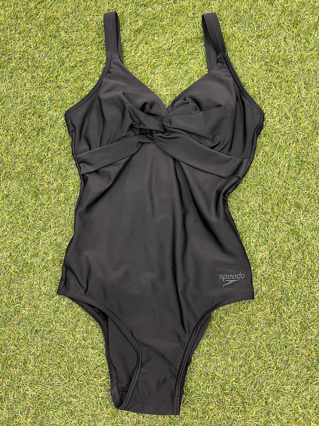 Front view of Speedo black one-piece swimsuit with twist front detail laid flat on grass