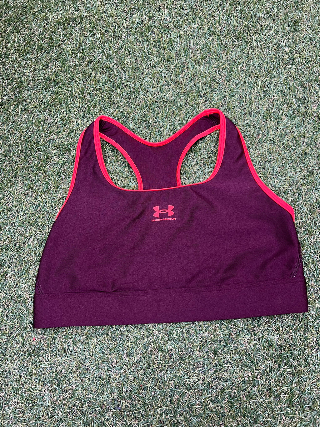 Front view of Under Armour sports bra in burgundy with pink trim laid flat on grass