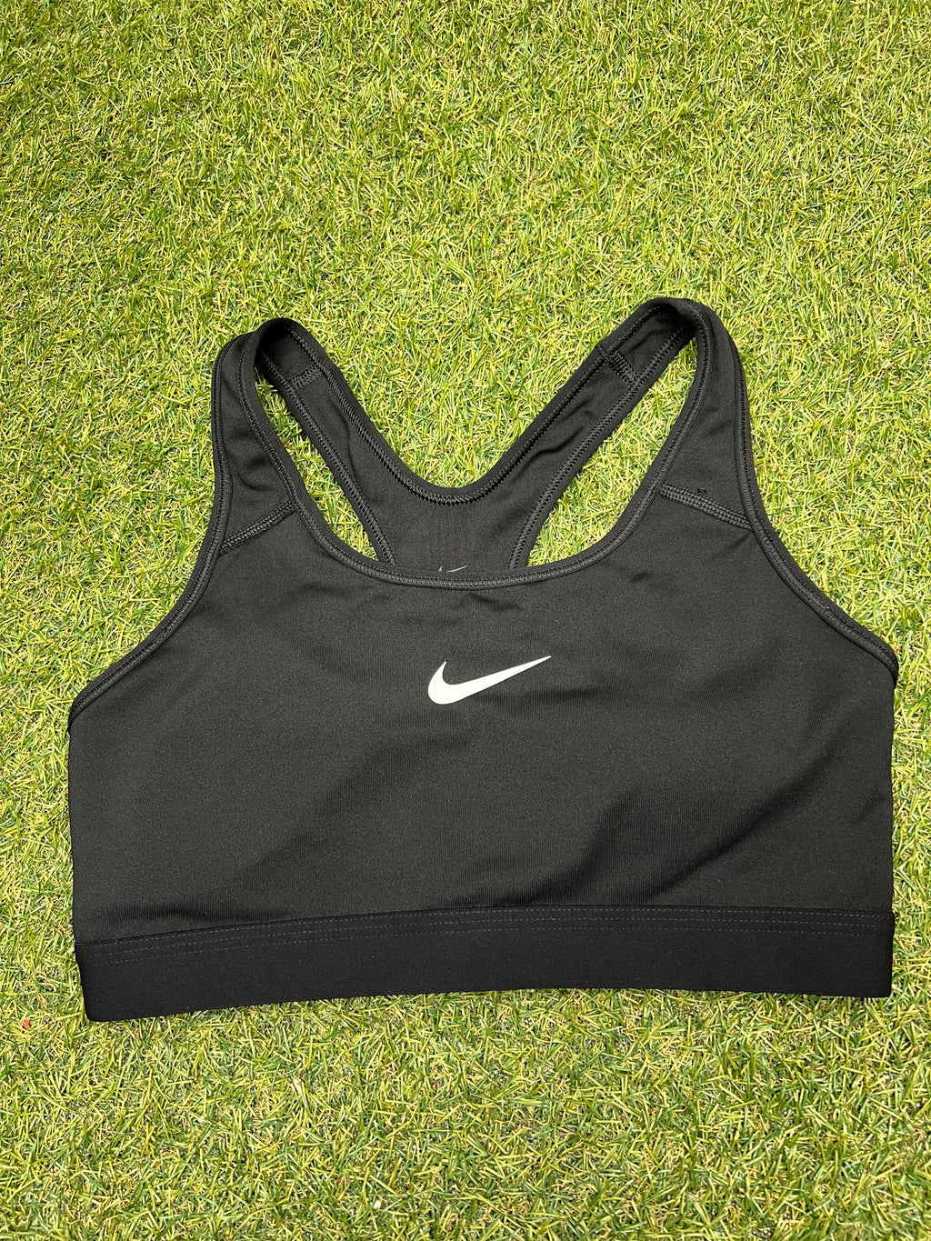 Front view of Nike black sports bra top laid on grass with white Nike swoosh