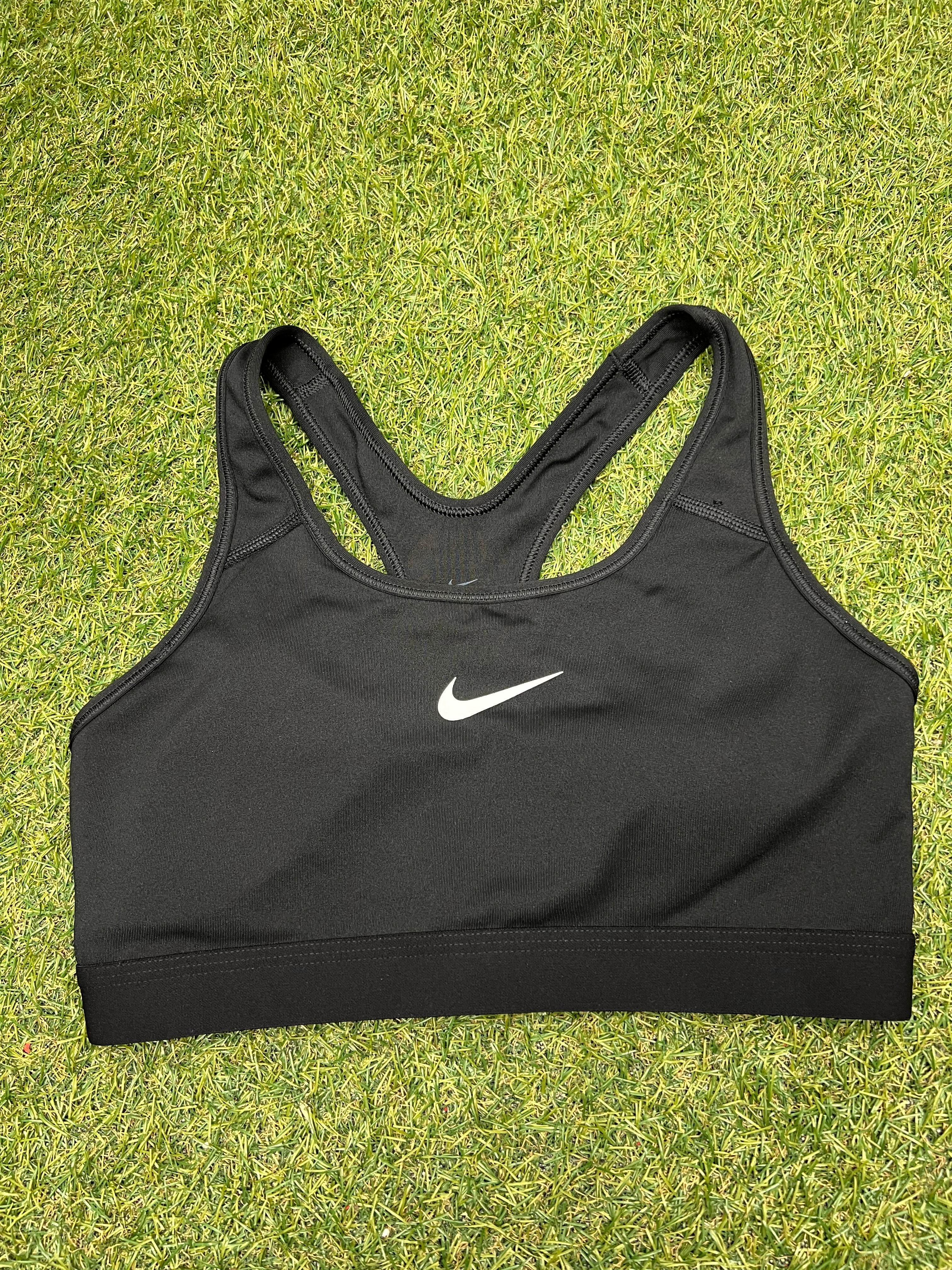 Front view of Nike black sports bra top laid on grass with white Nike swoosh