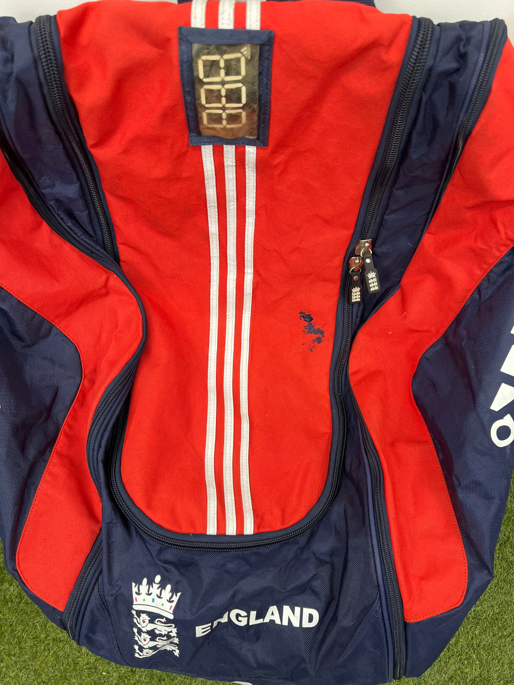 Close-up of top section of cricket bag showing red panel with white stripes and zip closure