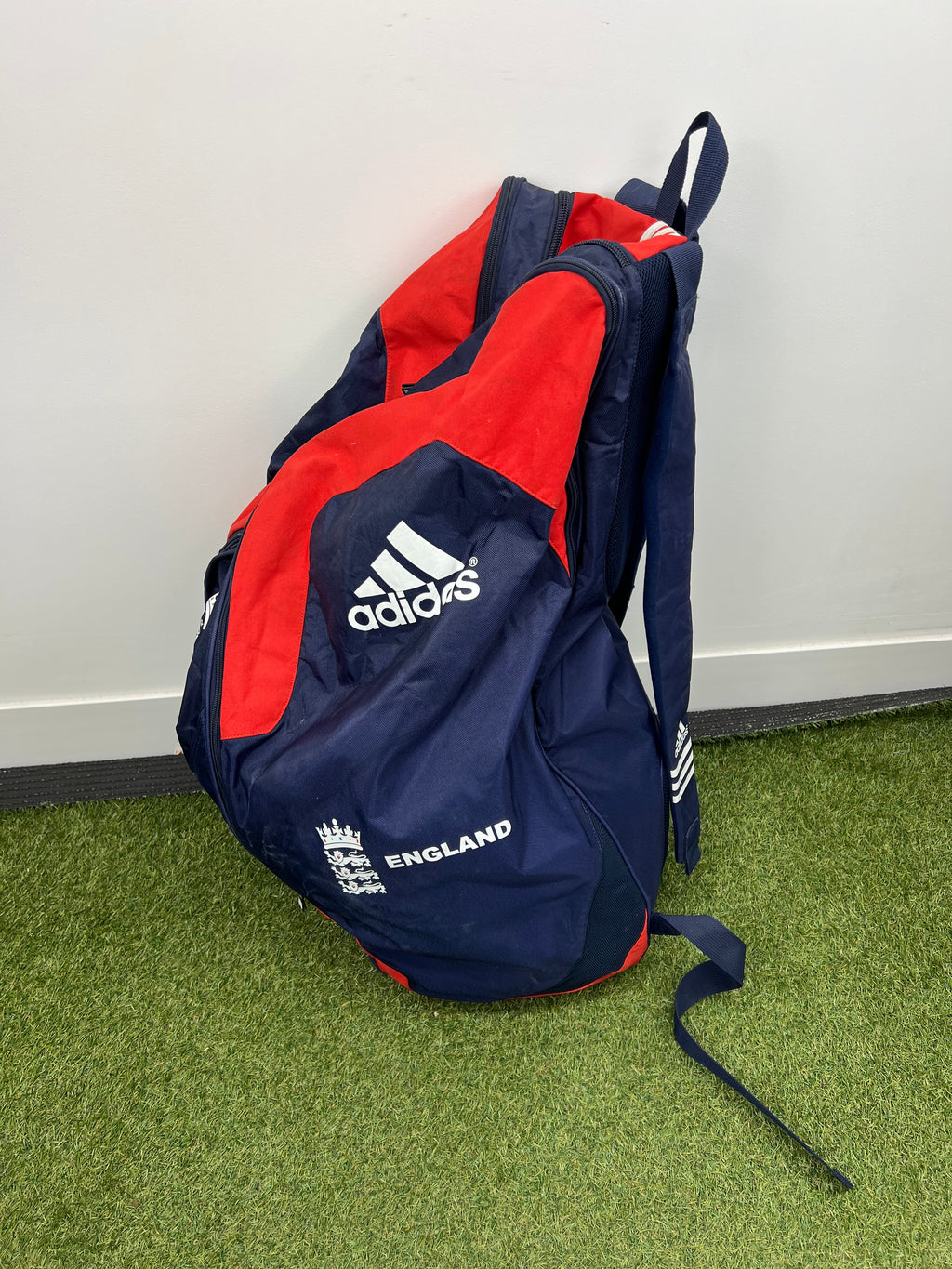 Side profile of cricket bag in red and navy, standing upright on artificial grass