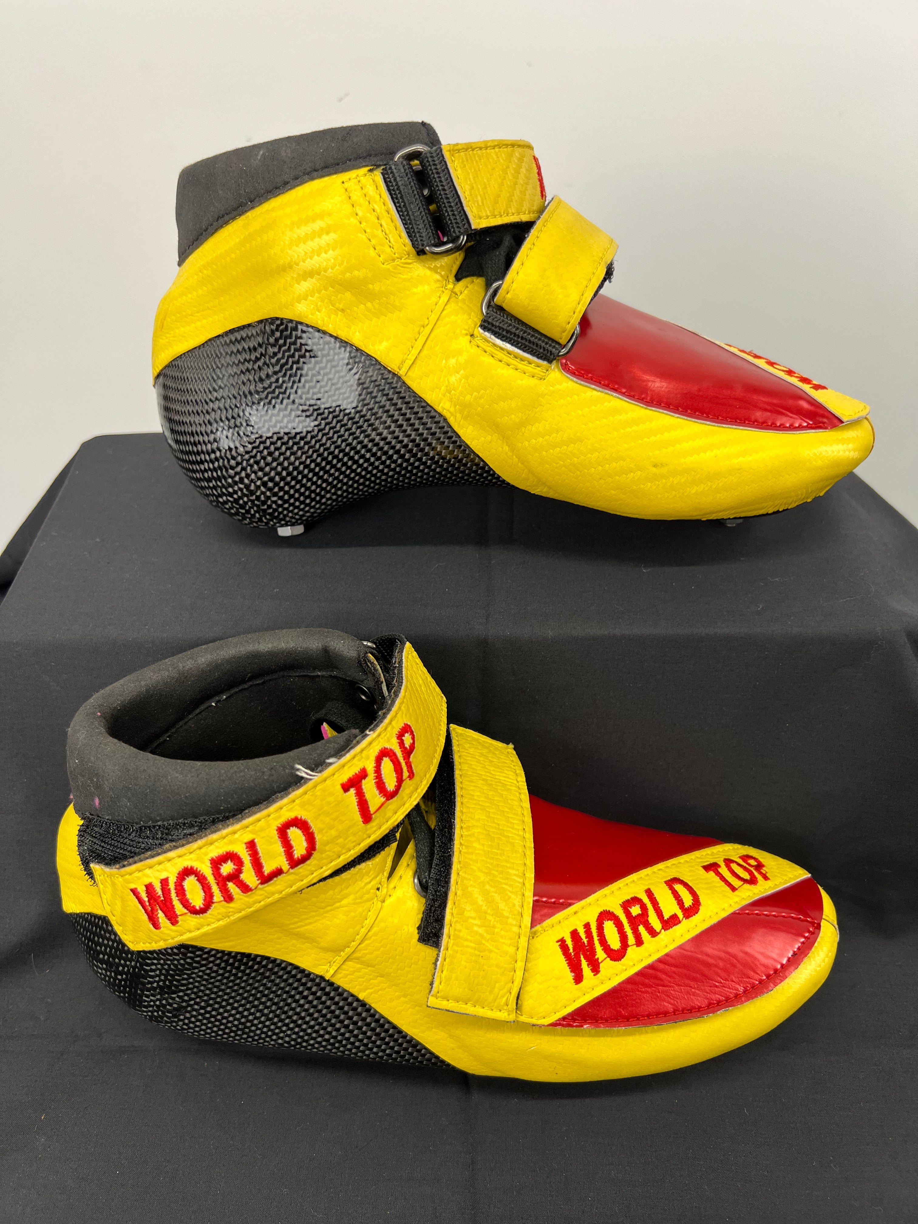 Side profile of a yellow and red World Top short track skate size 38 showing carbon fibre heel and two yellow straps with red lettering