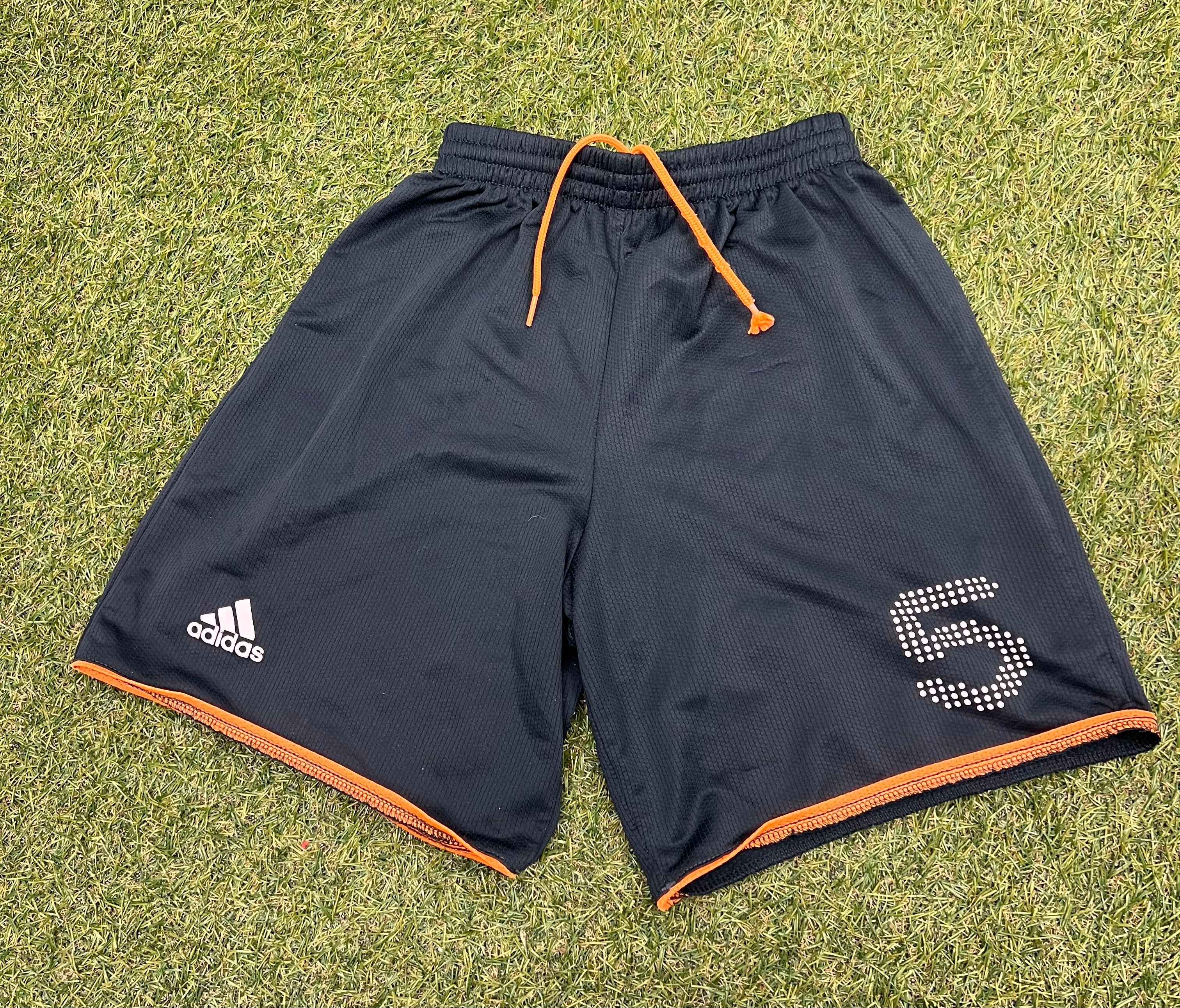 Black kids adidas shorts with orange trim and No 5 print on front, laid flat on grass