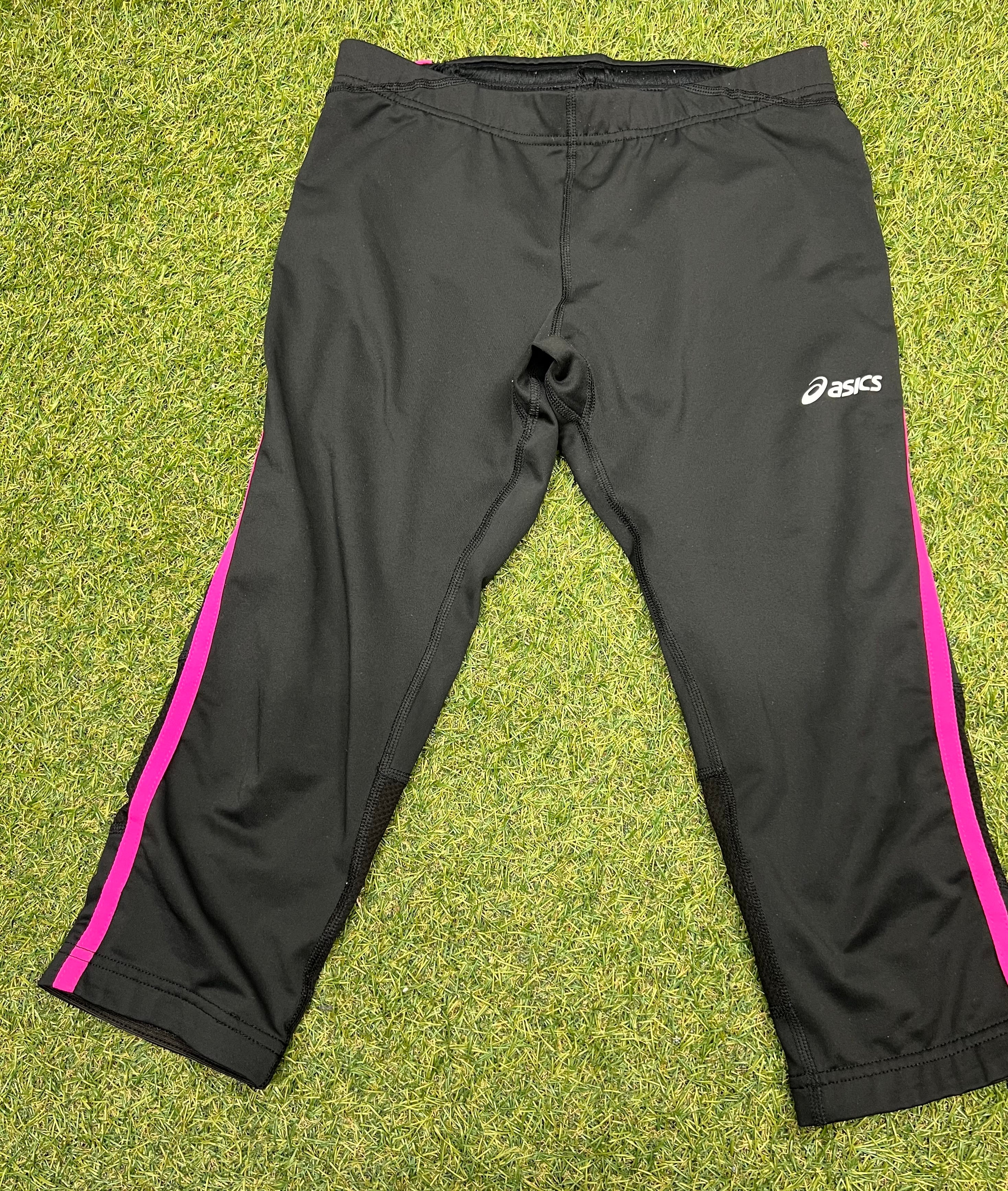 Front view of Asics black cropped leggings with pink side stripes laid flat on grass