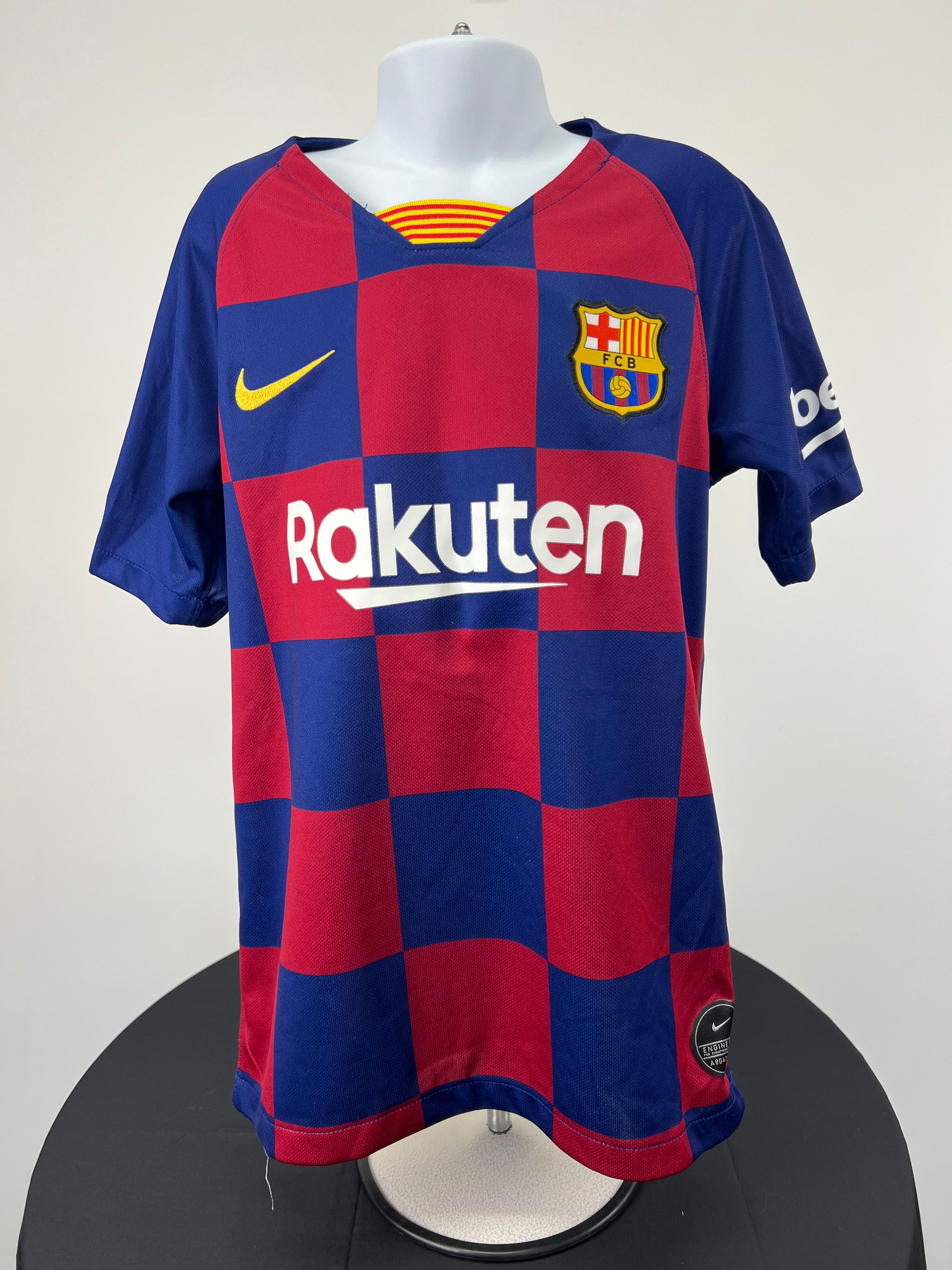 Front view of kids FC Barcelona football shirt with red and blue check pattern, Rakuten logo, Nike swoosh and FC Barcelona crest