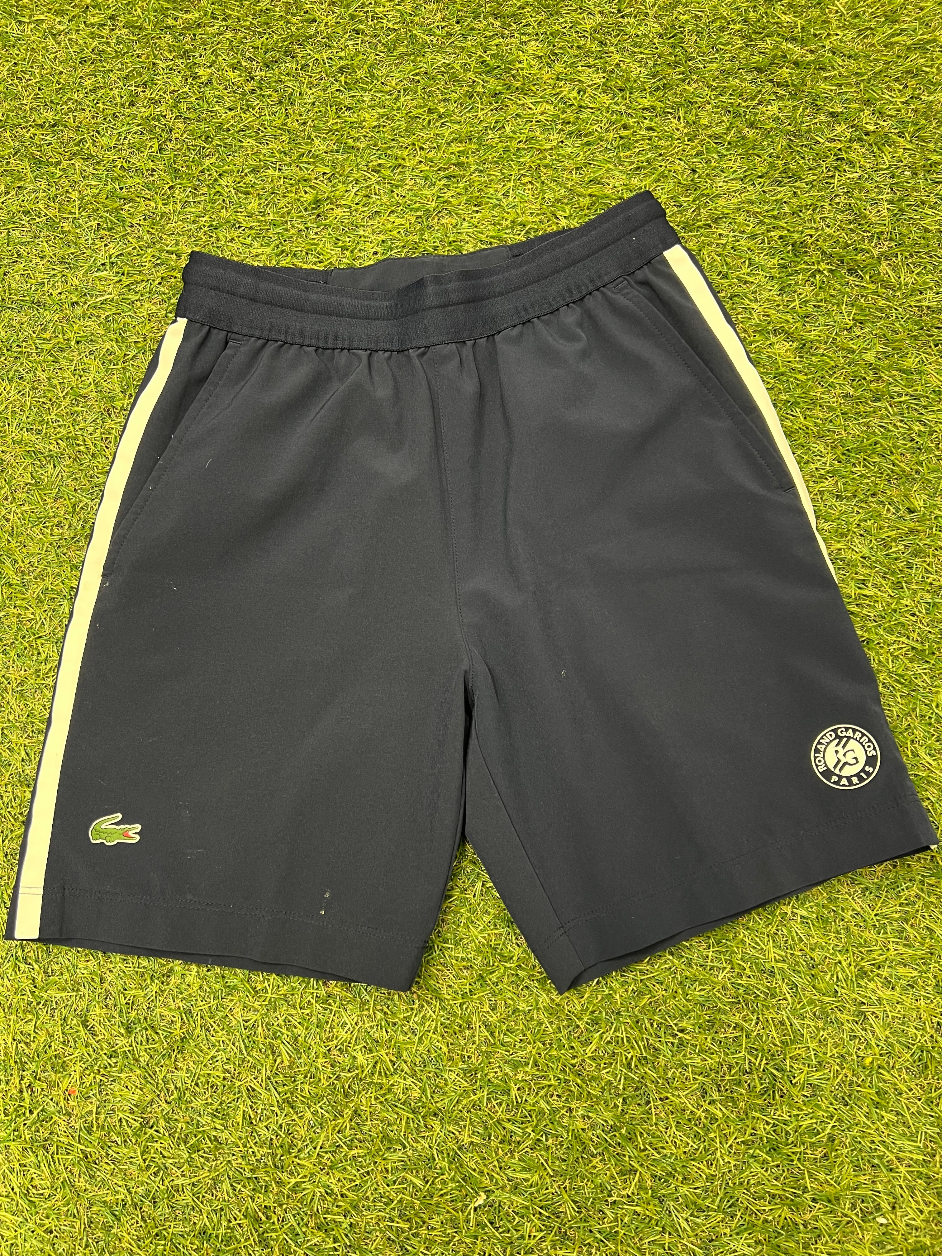 Front view of black Lacoste Roland Garros kids shorts with yellow side stripes on grass